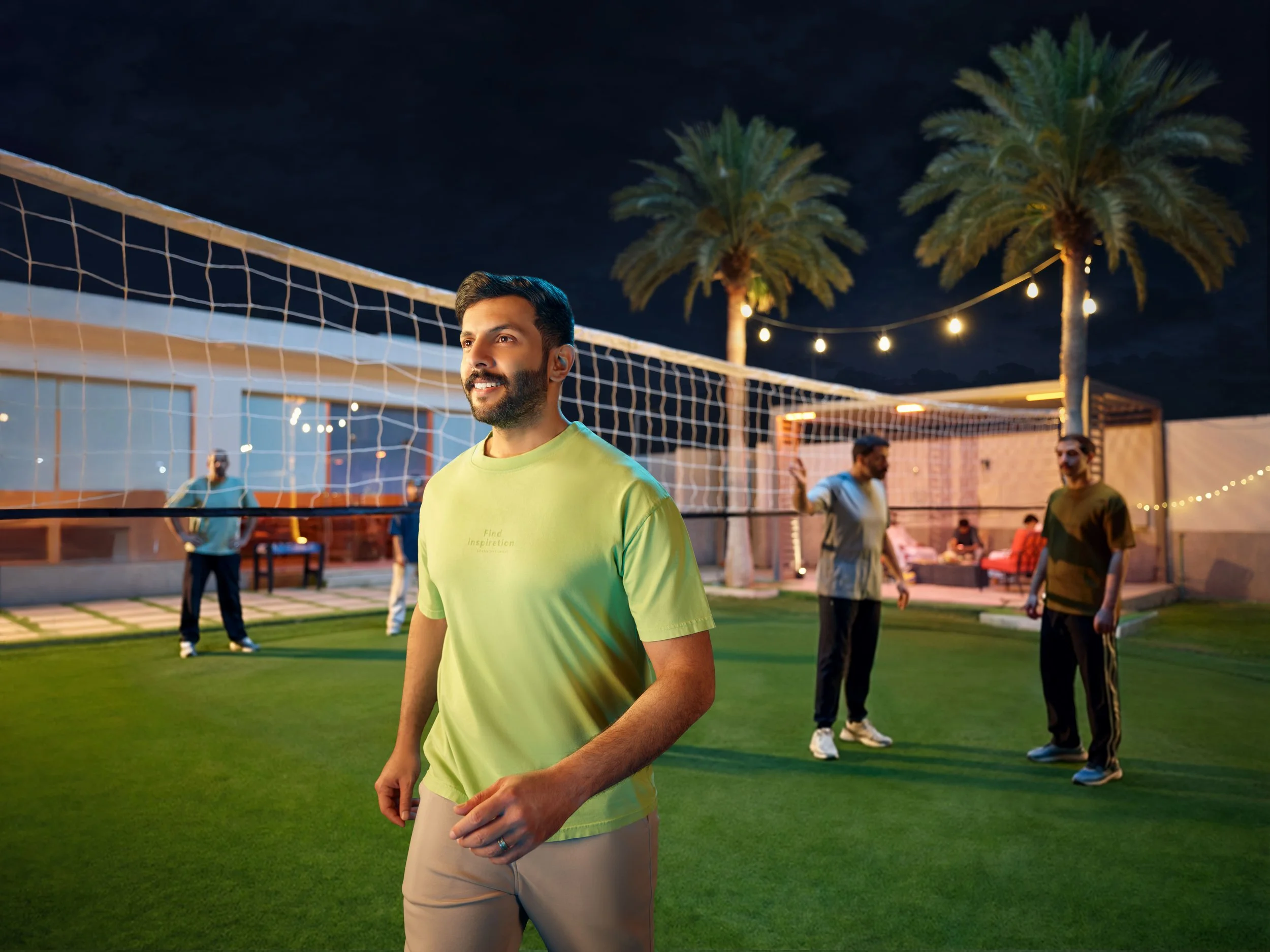 Man in lime green shirt standing on a volleyball court at night, with others in the background playing or socializing, illuminated by string lights and palm trees.