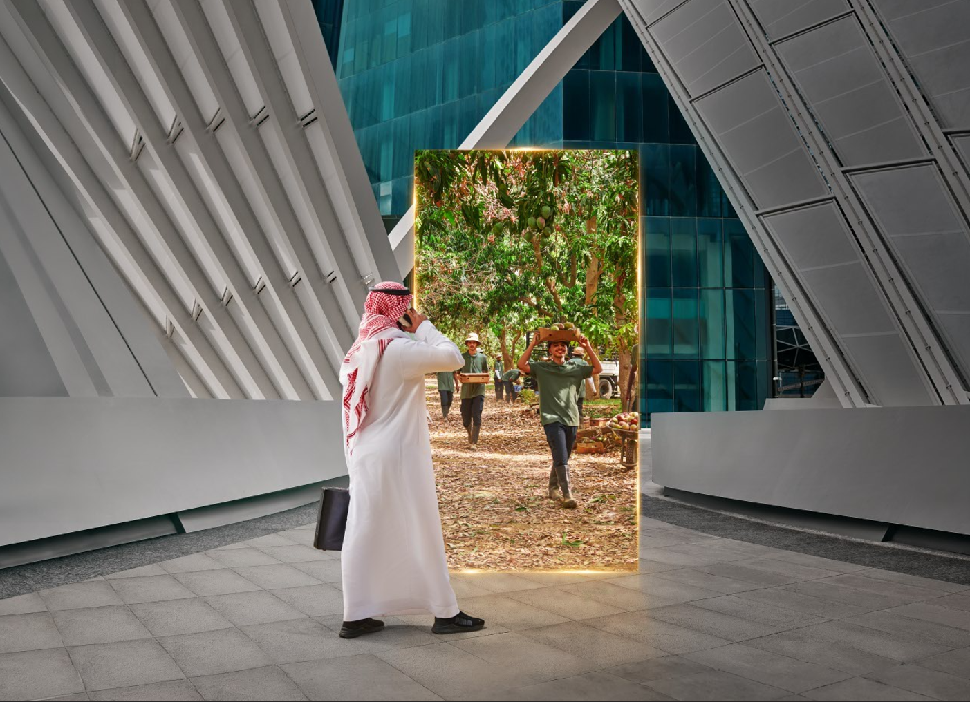 A man dressed in traditional Middle Eastern attire taking a photo with his phone of a scene behind a mirror inside a modern building. The scene behind the mirror shows people harvesting fruit in an orchard.