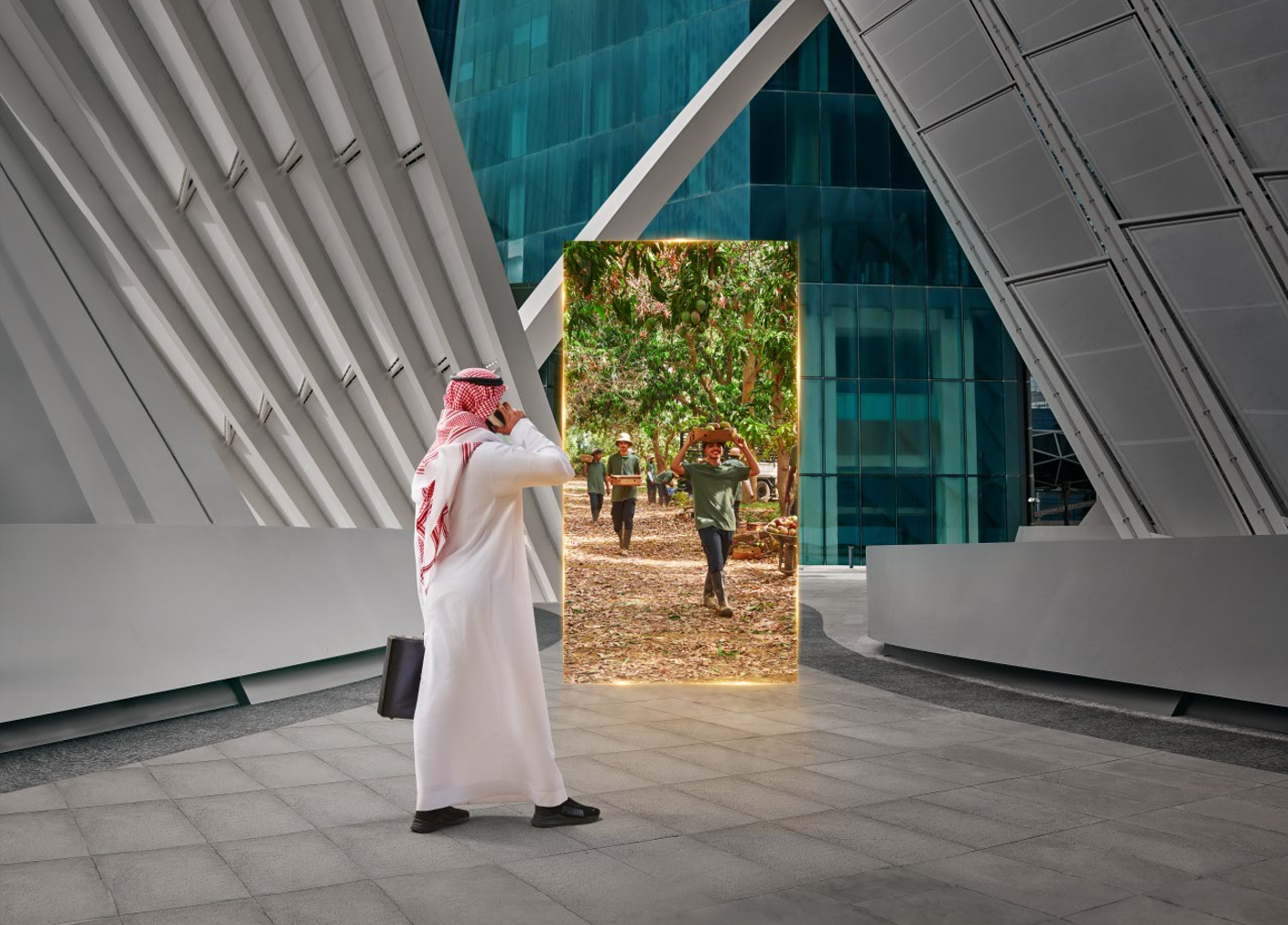 A man in traditional Middle Eastern attire taking a photo of a large vertical screen displaying a scene of people working outdoors under trees, with modern architecture around him.
