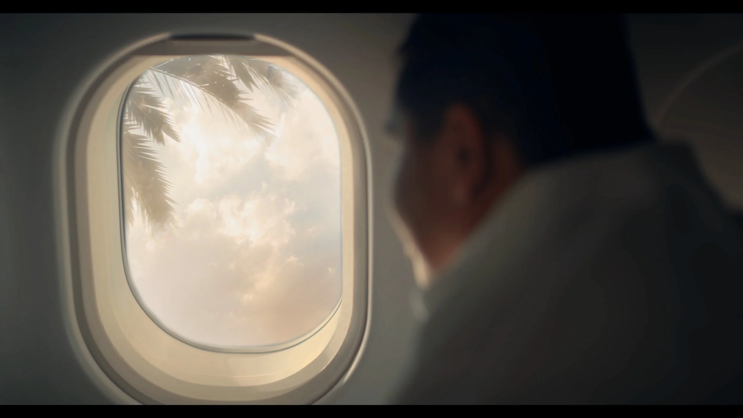 A person wearing glasses and a face mask looking out of an airplane window at clouds and palm trees.