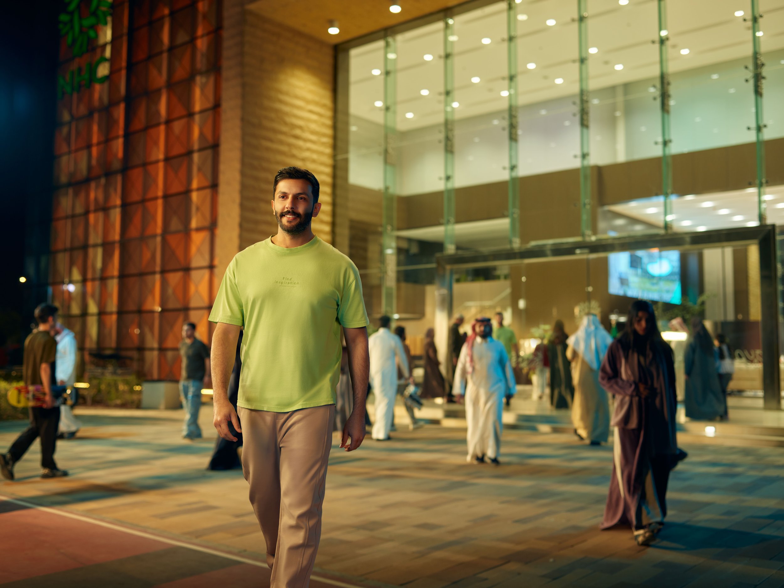 A young man with a beard wearing a green T-shirt and beige pants walking outside a modern building at night, with several people in traditional Middle Eastern attire in the background.