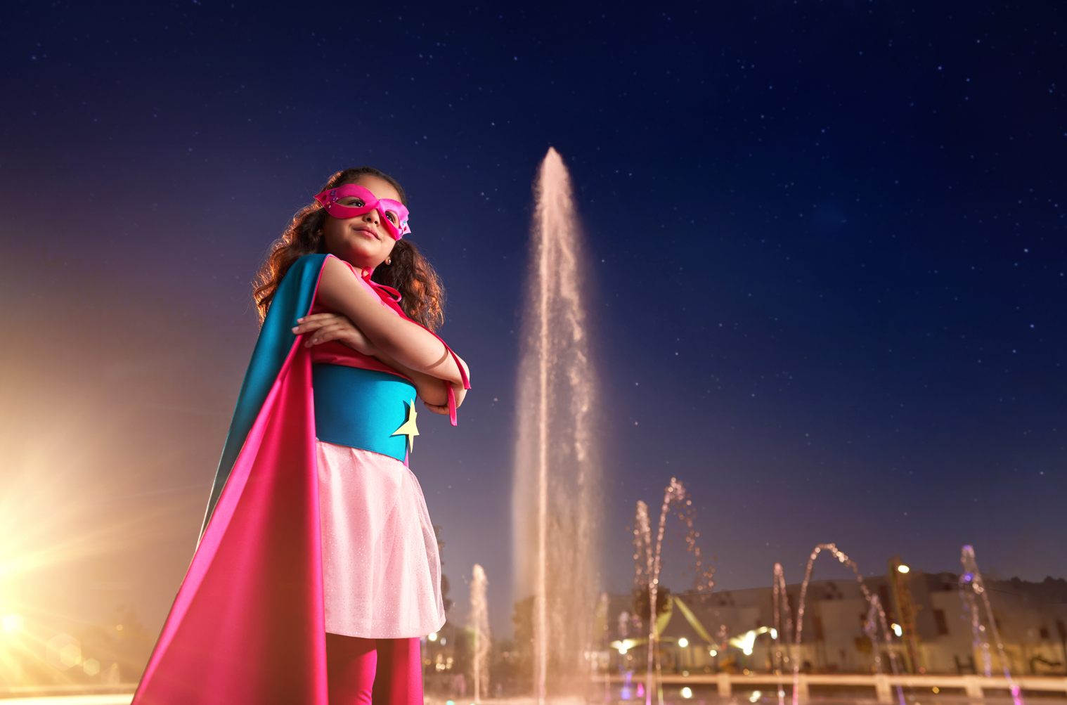 Young girl dressed as a superhero with a pink mask and a cape, standing with arms crossed in front of a fountain at night, with the water fountain jet shooting upward and city lights in the background.