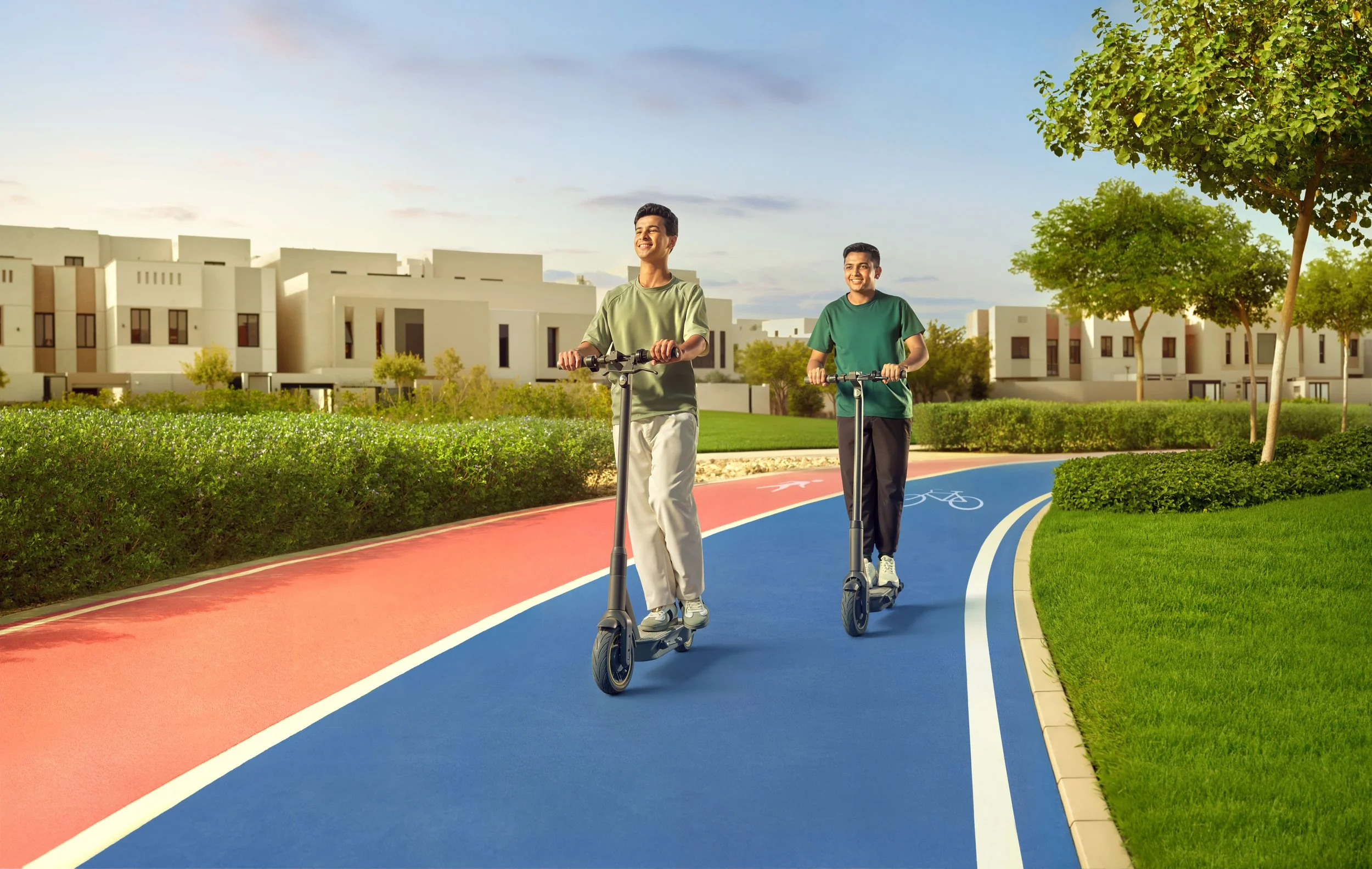 Two young men riding electric scooters on a colorful, curved bike path in a park with green trees and modern white buildings in the background.