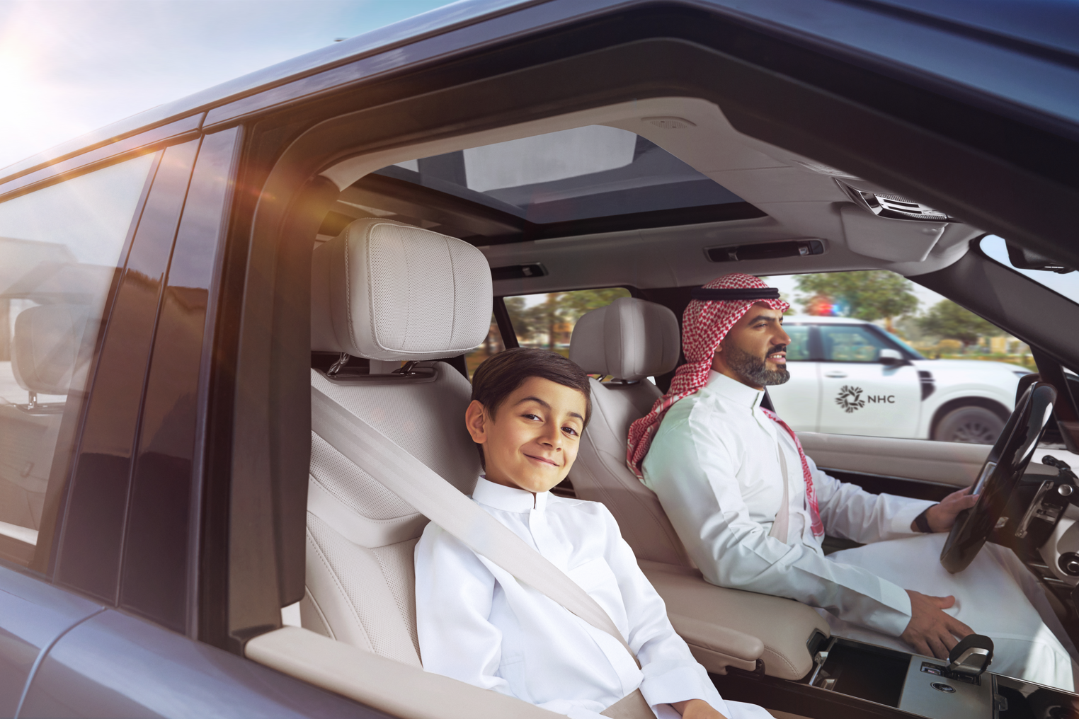 An adult male in traditional Middle Eastern attire, including a red and white keffiyeh, driving a car with a young boy in traditional white clothing sitting in the passenger seat. The boy is smiling and wearing a seatbelt, while the man is focused on