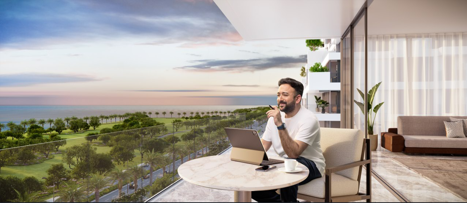 Man sitting at a marble table with a tablet and coffee cup on balcony overlooking a park and ocean during sunset.