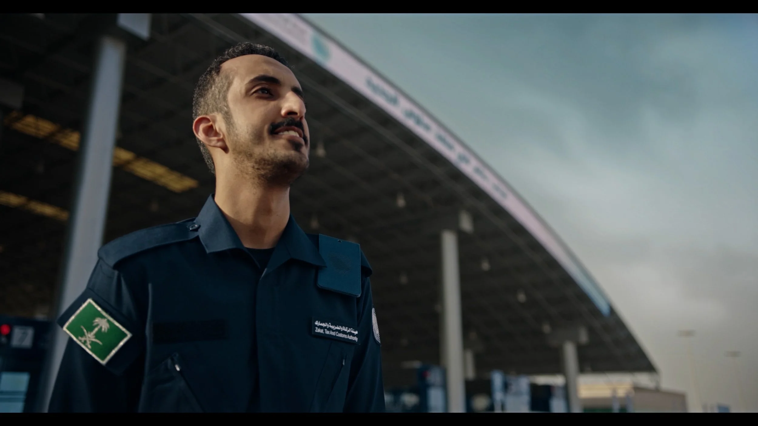 A man in a TSA uniform at an airport, with a large curved roof structure above and some airport signs in the background.