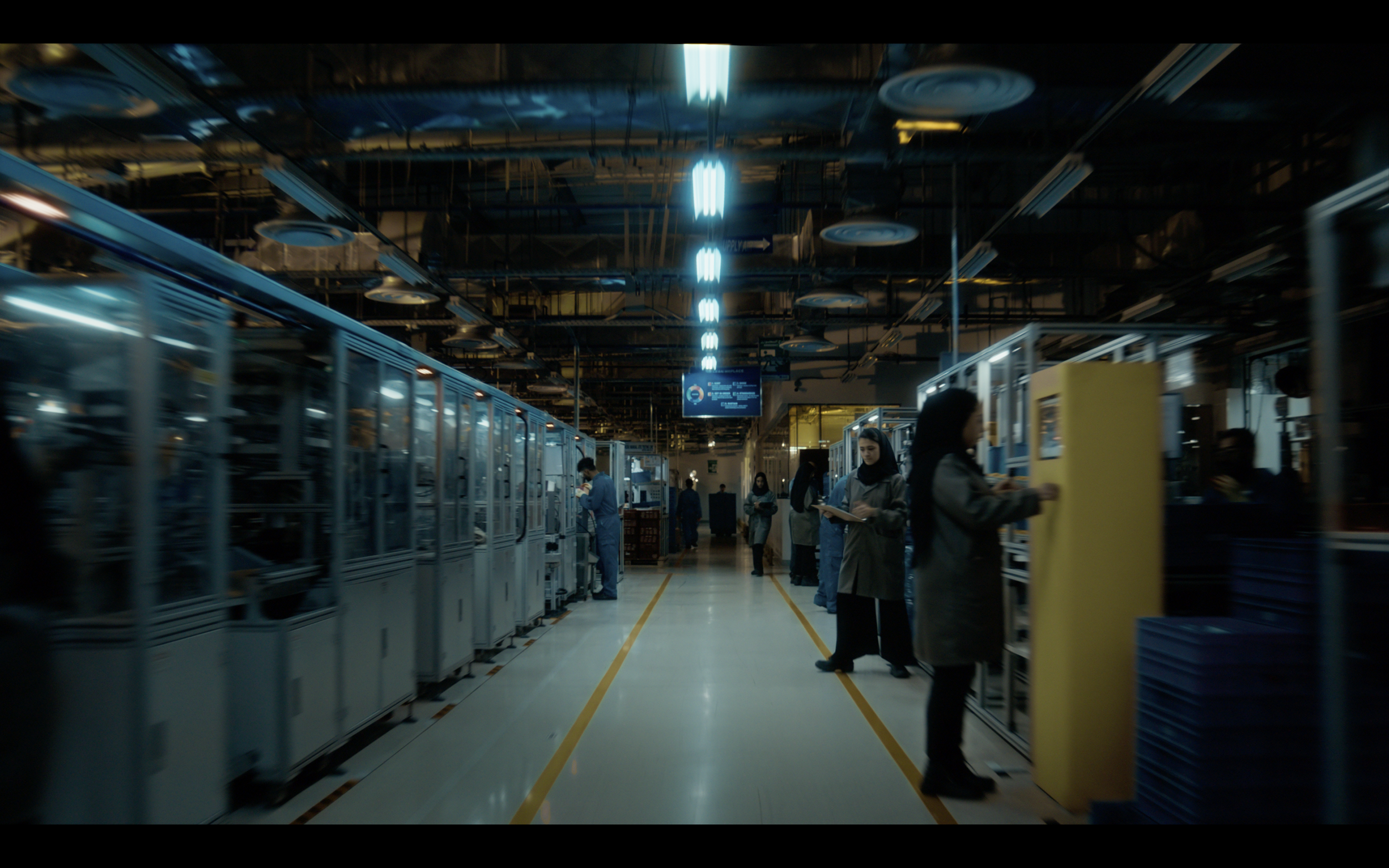 Inside a technology manufacturing facility with employees working on assembly lines and machines under fluorescent lighting.