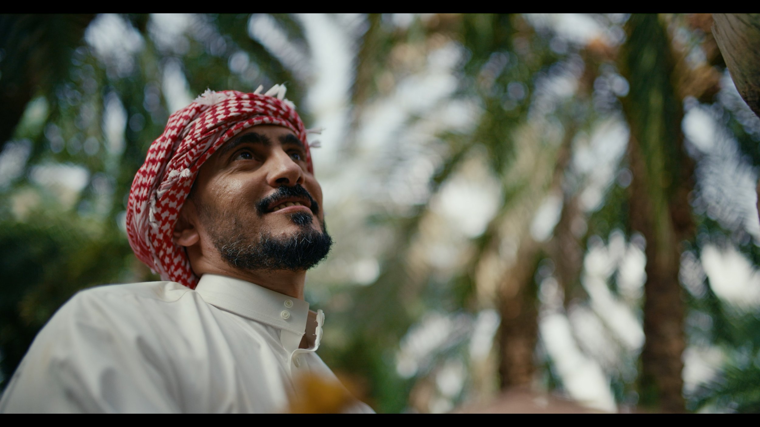 A man wearing a traditional Middle Eastern outfit and a red-and-white checkered keffiyeh looking upward in a lush outdoor setting.