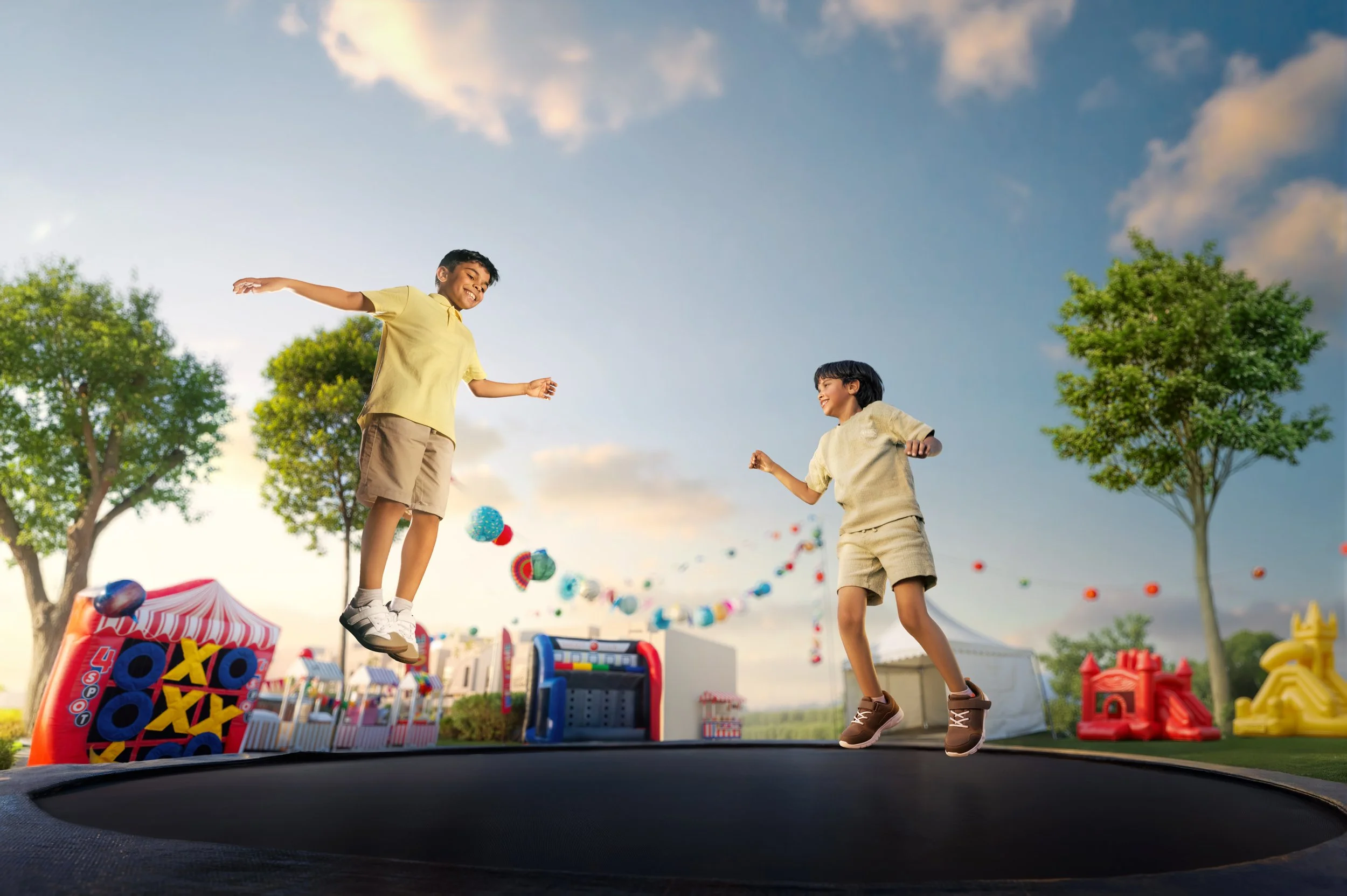 Two young boys jumping on a trampoline in a colorful outdoor fair or park with trees, tents, and play equipment in the background.