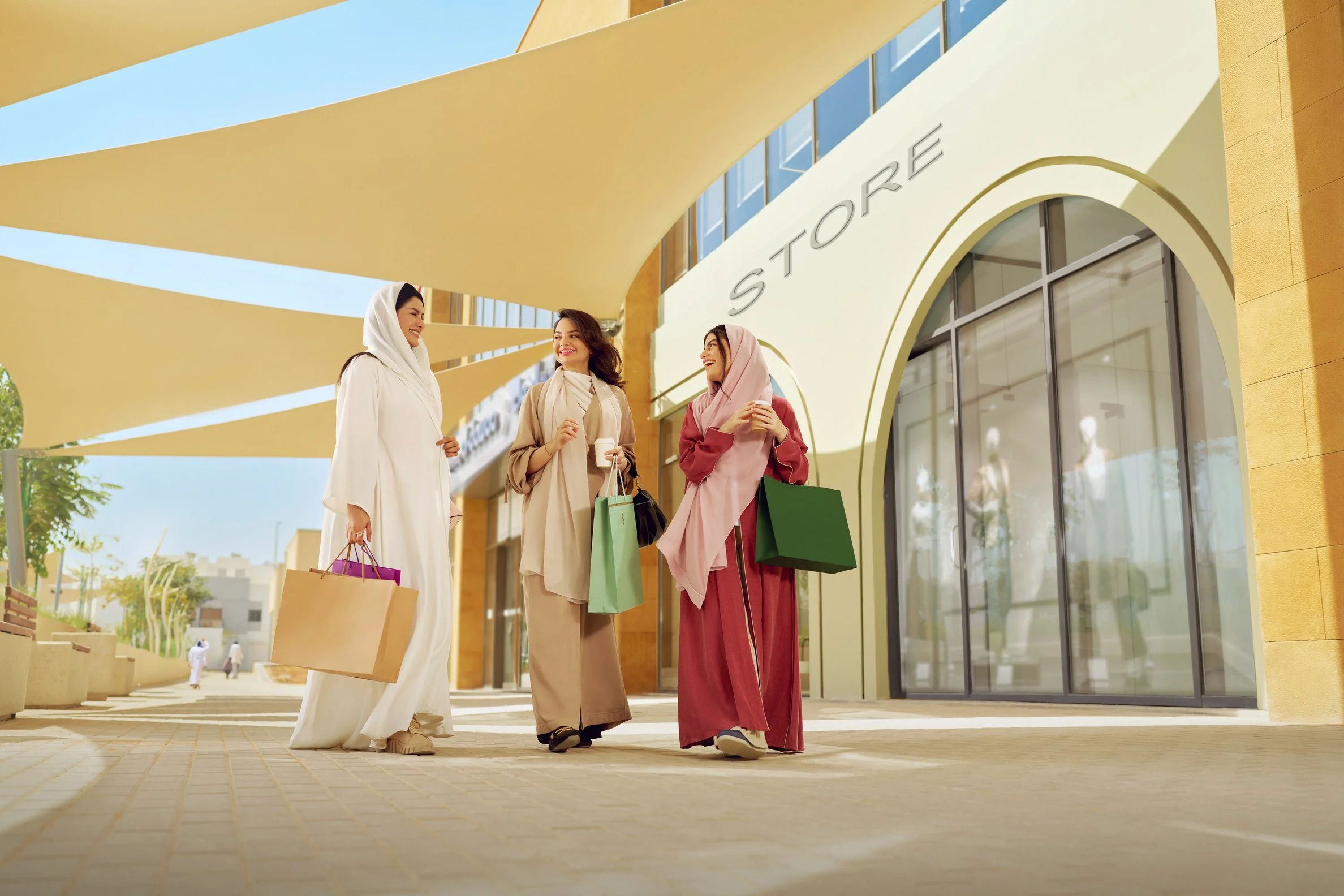 Three women in hijabs with shopping bags and cups of coffee, smiling and chatting outside a shopping mall with a yellow building and canopy.