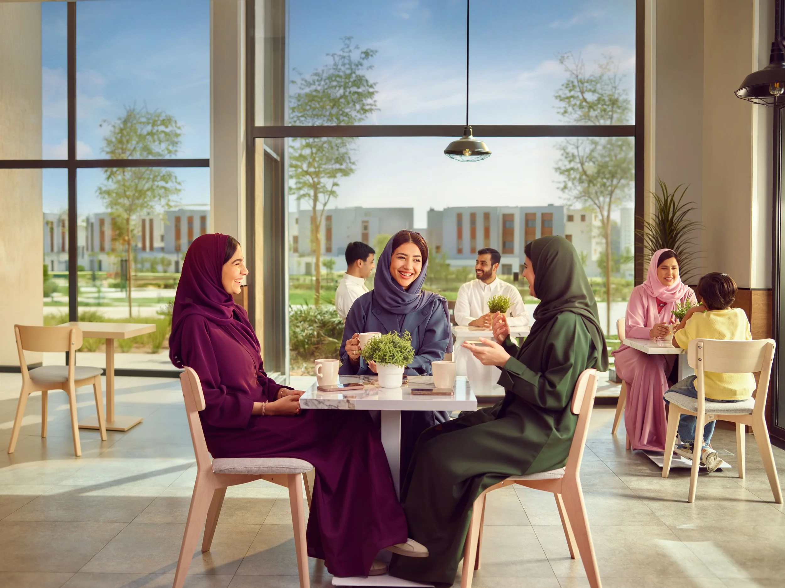 Group of women and kids enjoying coffee in a modern café during daylight, with large windows and a view of trees and buildings outside.