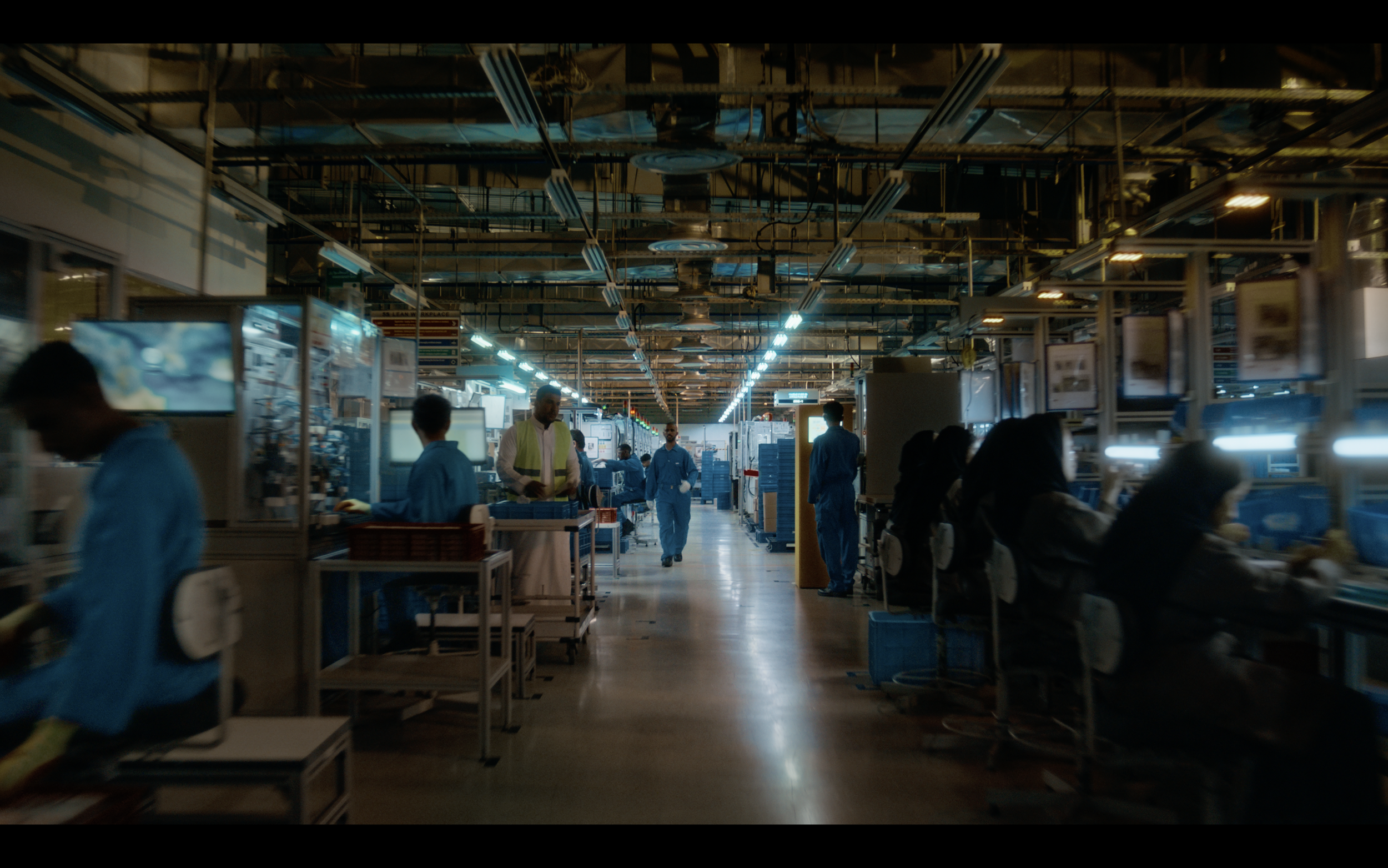 Workers in blue uniforms working on electronic devices in a manufacturing factory with overhead lighting and exposed ceiling pipes.