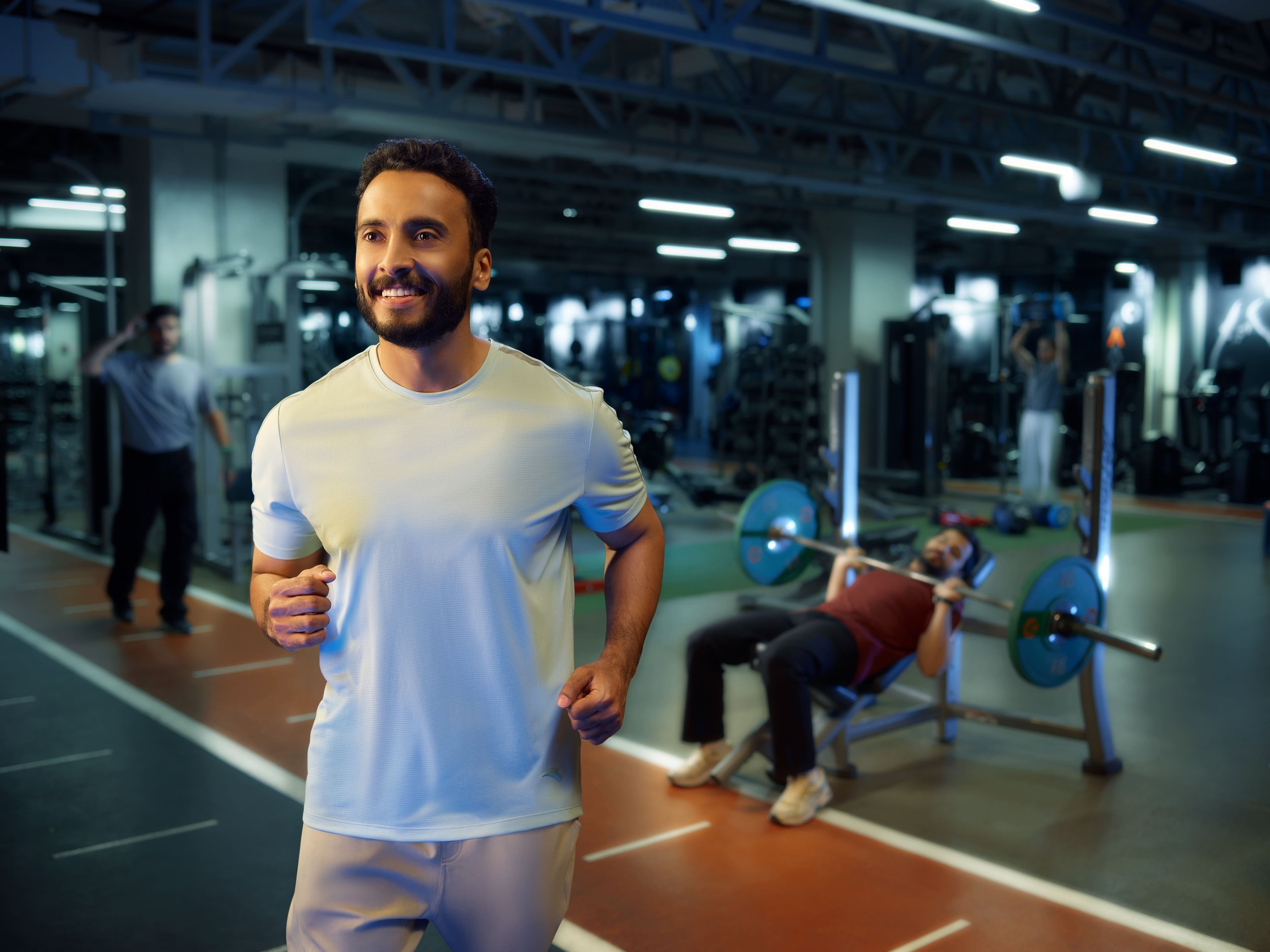 A smiling man jogging inside a gym with other people exercising in the background, including a woman lifting weights on a bench.