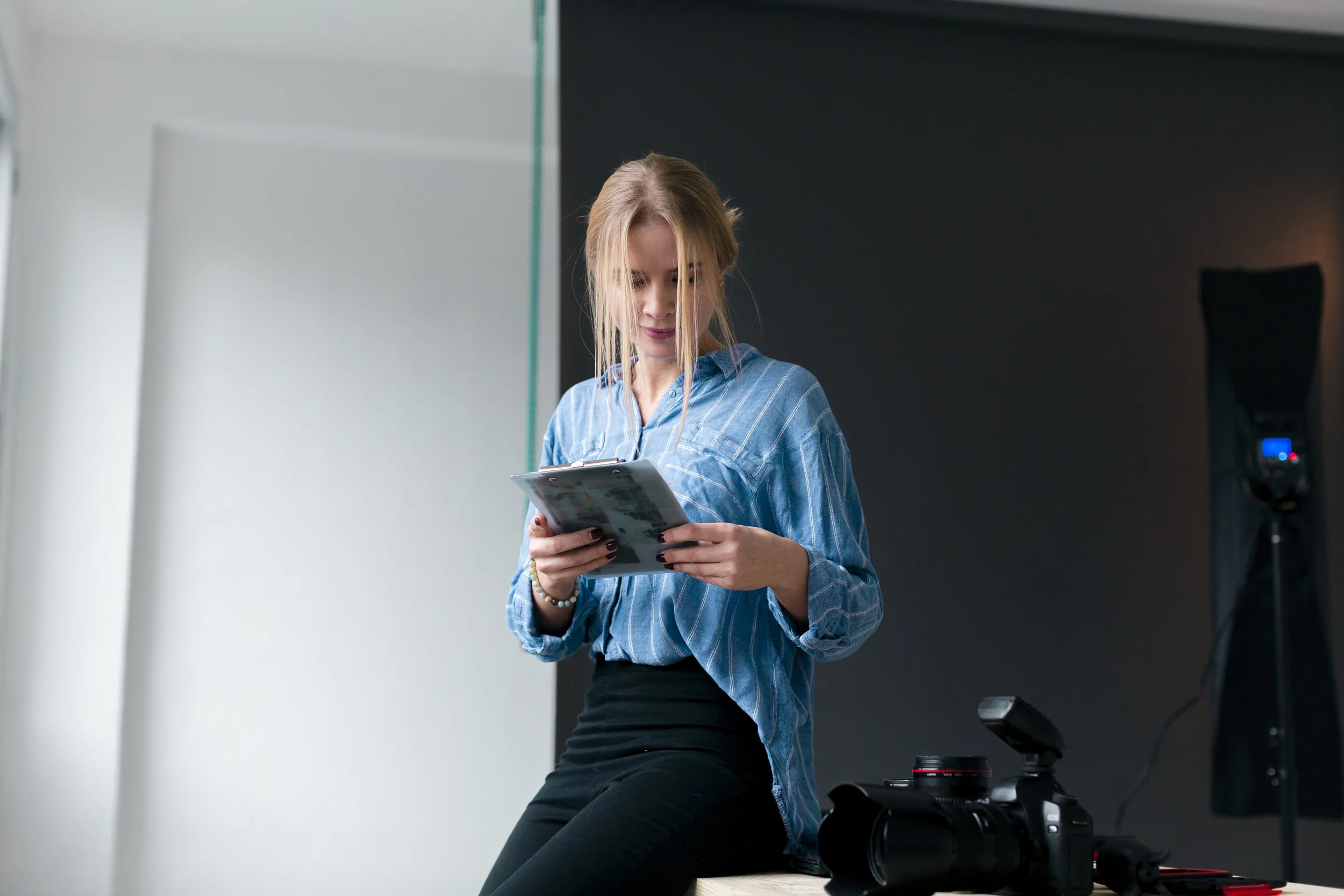 A woman with blonde hair in a blue plaid shirt and black pants sitting on a table, looking at a tablet, with a camera nearby in a photography studio.