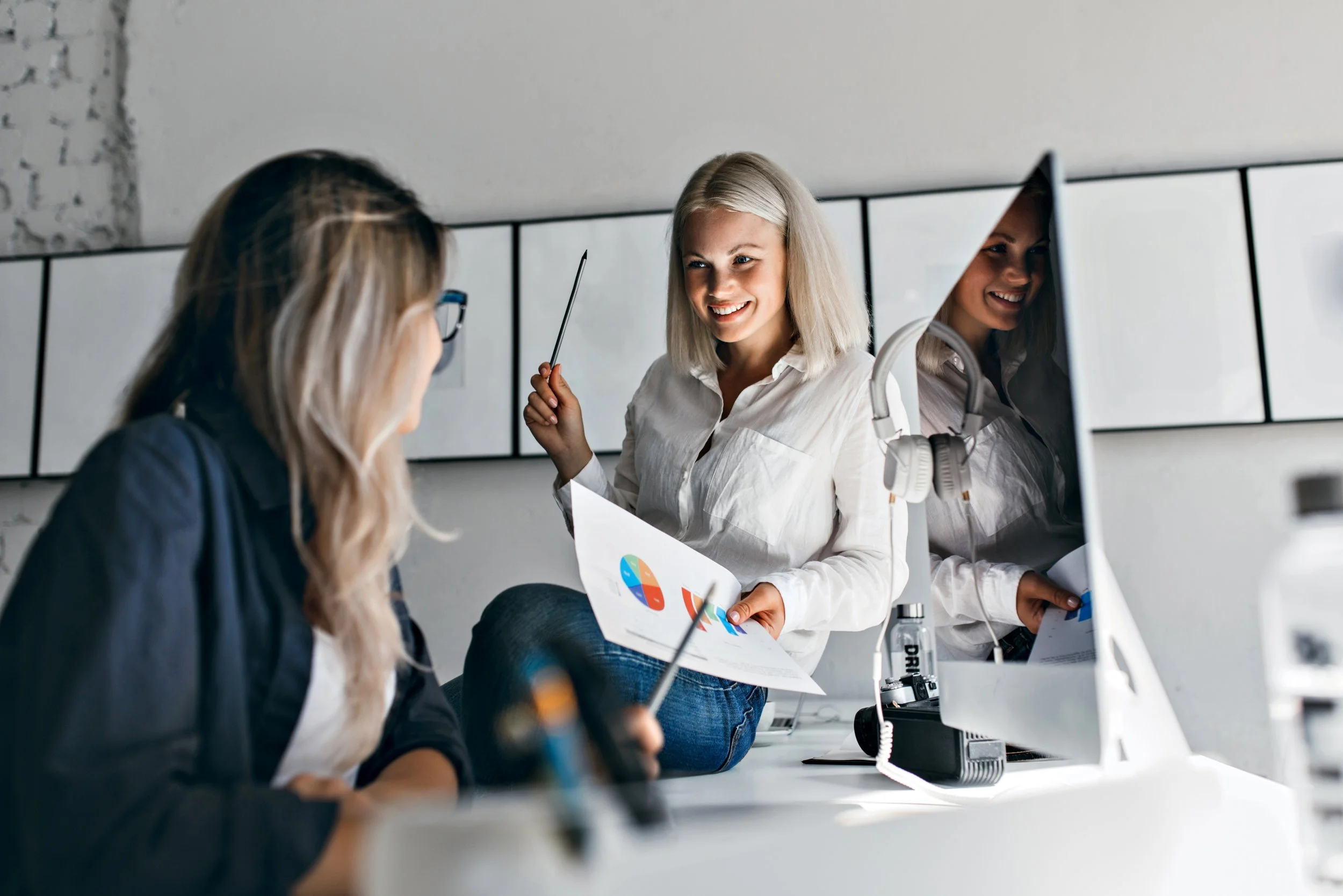 Three women in an office, one smiling and holding a document with pie charts, engaged in a discussion.