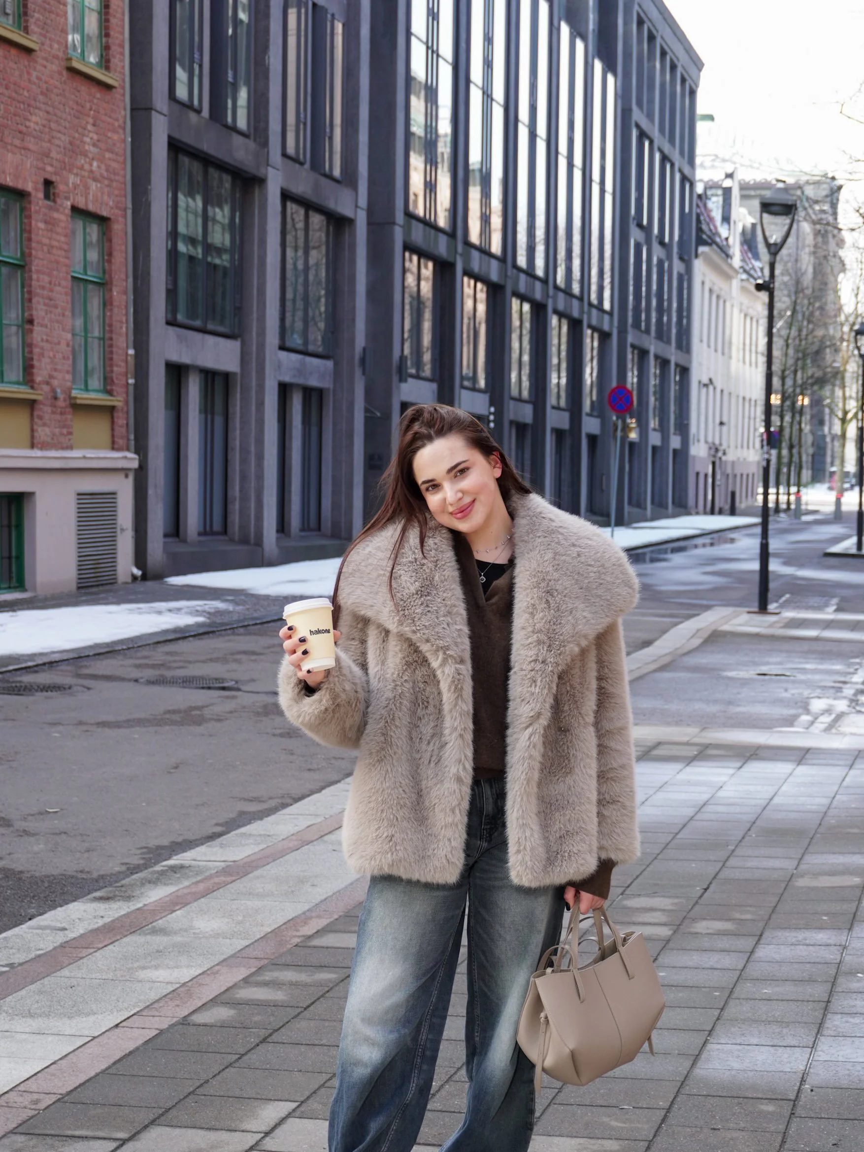 A woman in a beige fur coat and jeans is standing on a city sidewalk holding a coffee cup and a beige handbag, smiling at the camera.