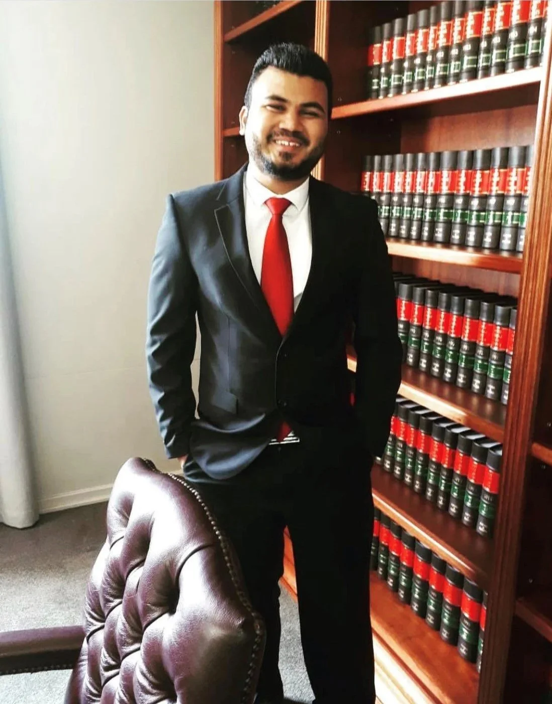 A man in a black suit, white shirt, and red tie standing in a law office or library. He is smiling and has his hands in his pockets, with a bookshelf filled with legal books in the background.