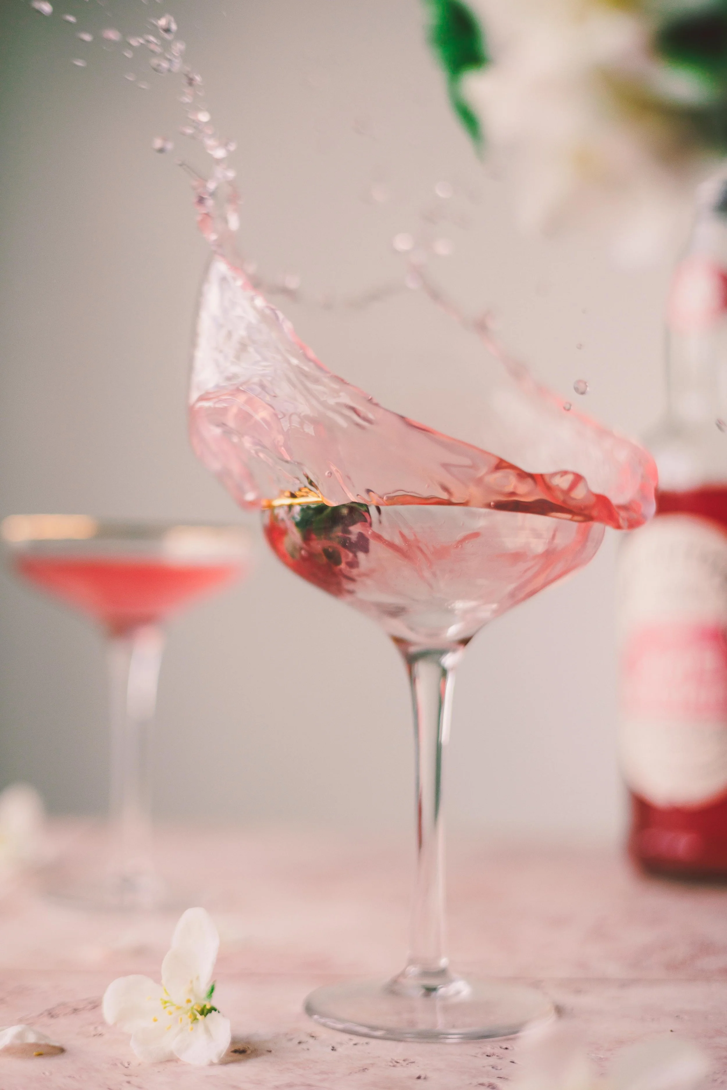 A pink cocktail splash in a martini glass, with another similar glass in the background and a blurred bottle on the right, on a pink marble surface with white flowers.