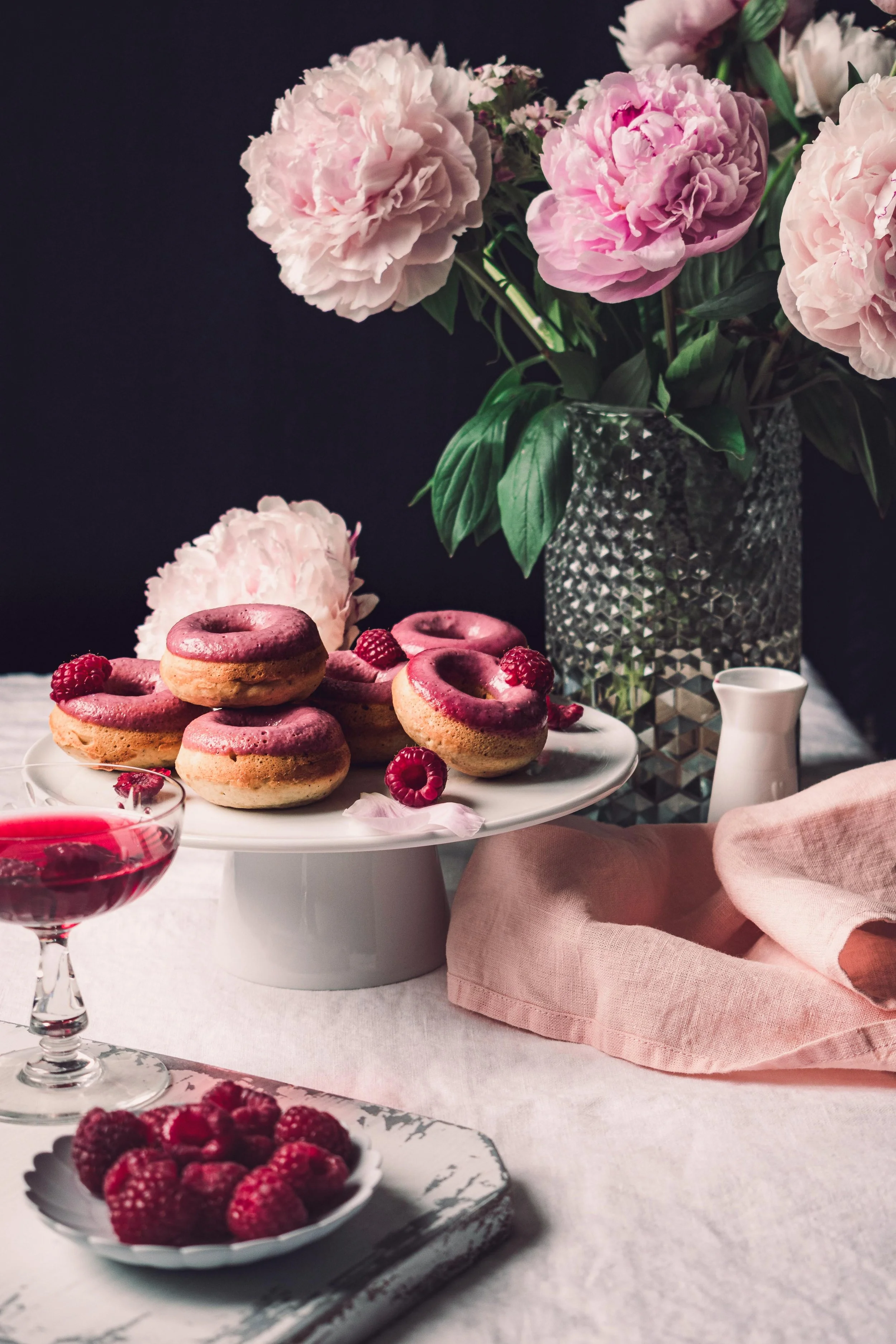 Pink and purple donuts with raspberry toppings on a white cake stand, surrounded by fresh raspberries, a glass of raspberry drink, a vase with pink peonies, a small white pitcher, and a pink napkin on a table.
