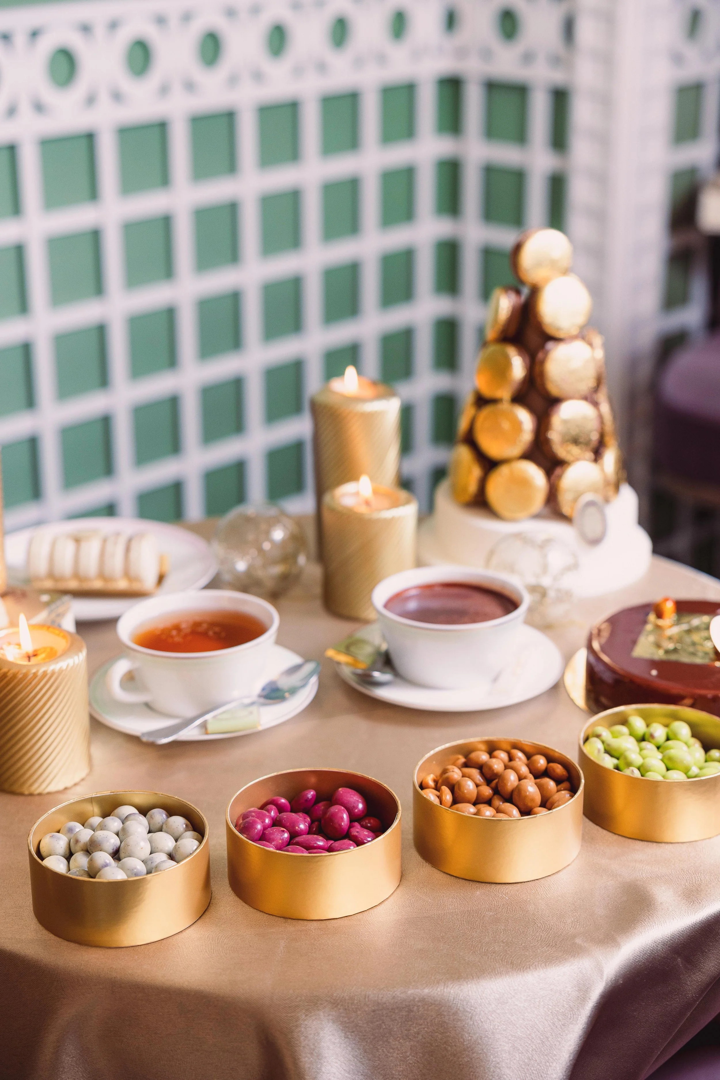 A decorated table with chocolates, teacups, candles, and a gold macaron tower for a celebration.