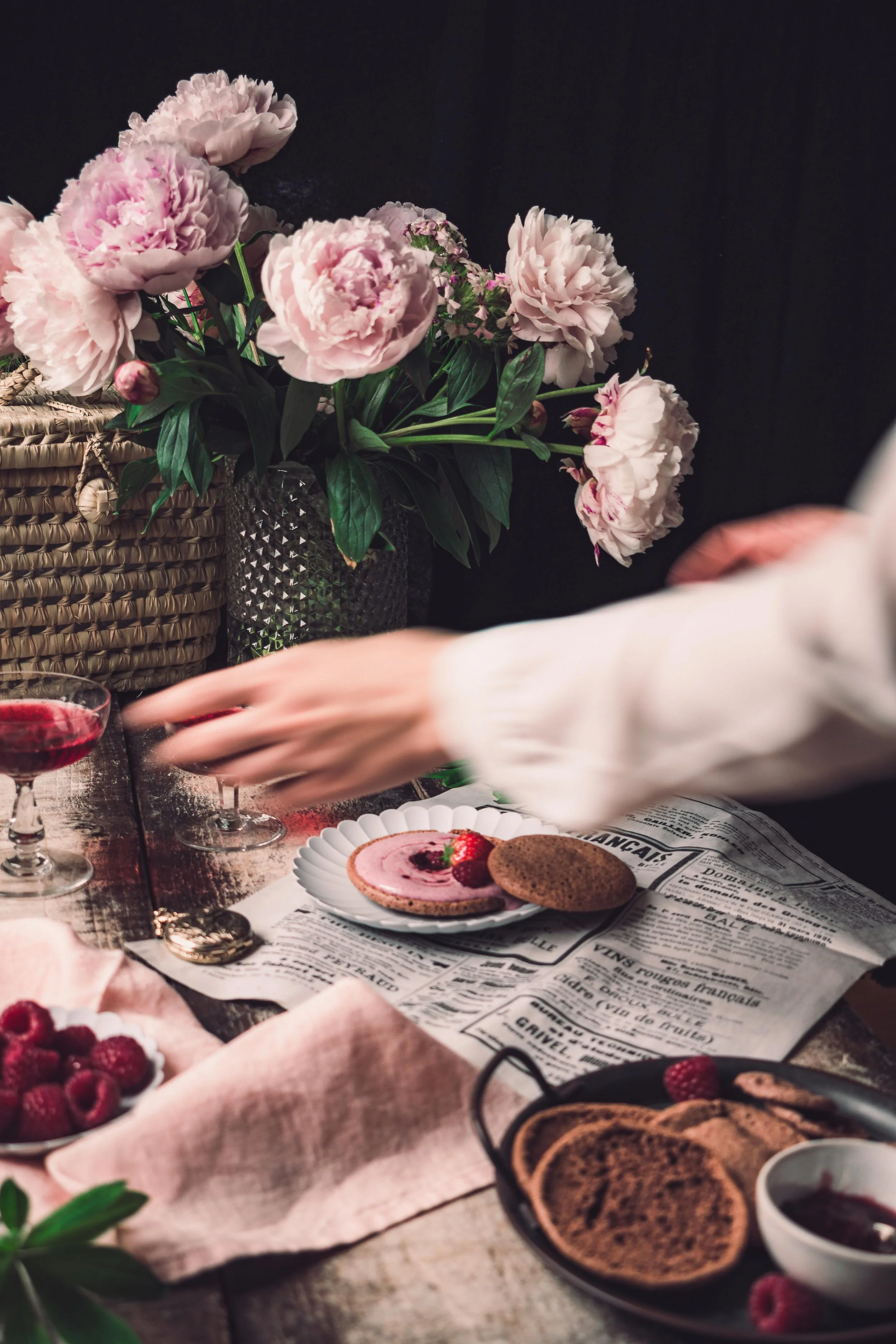 A table with pink peony flowers in a vase, a plate with raspberry-flavored mousse and a cookie, raspberries in a small bowl, and a cup of berry sauce, with a person's hand reaching for a glass of red wine.