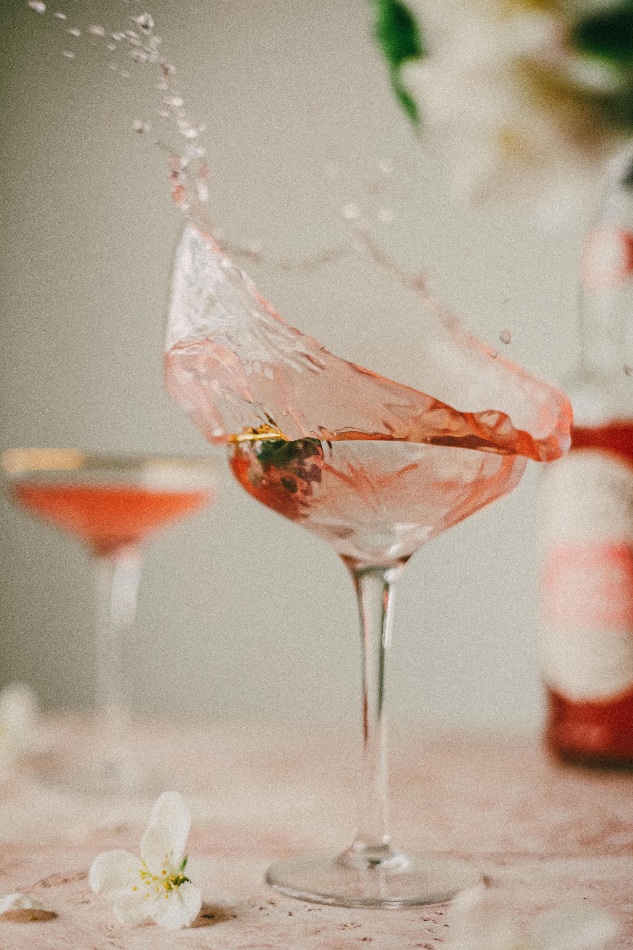 A pink cocktail in a margarita glass with a splash of liquid frozen mid-air, garnished with a small green leaf, on a soft pink surface with white flowers and a blurred red bottle in the background.