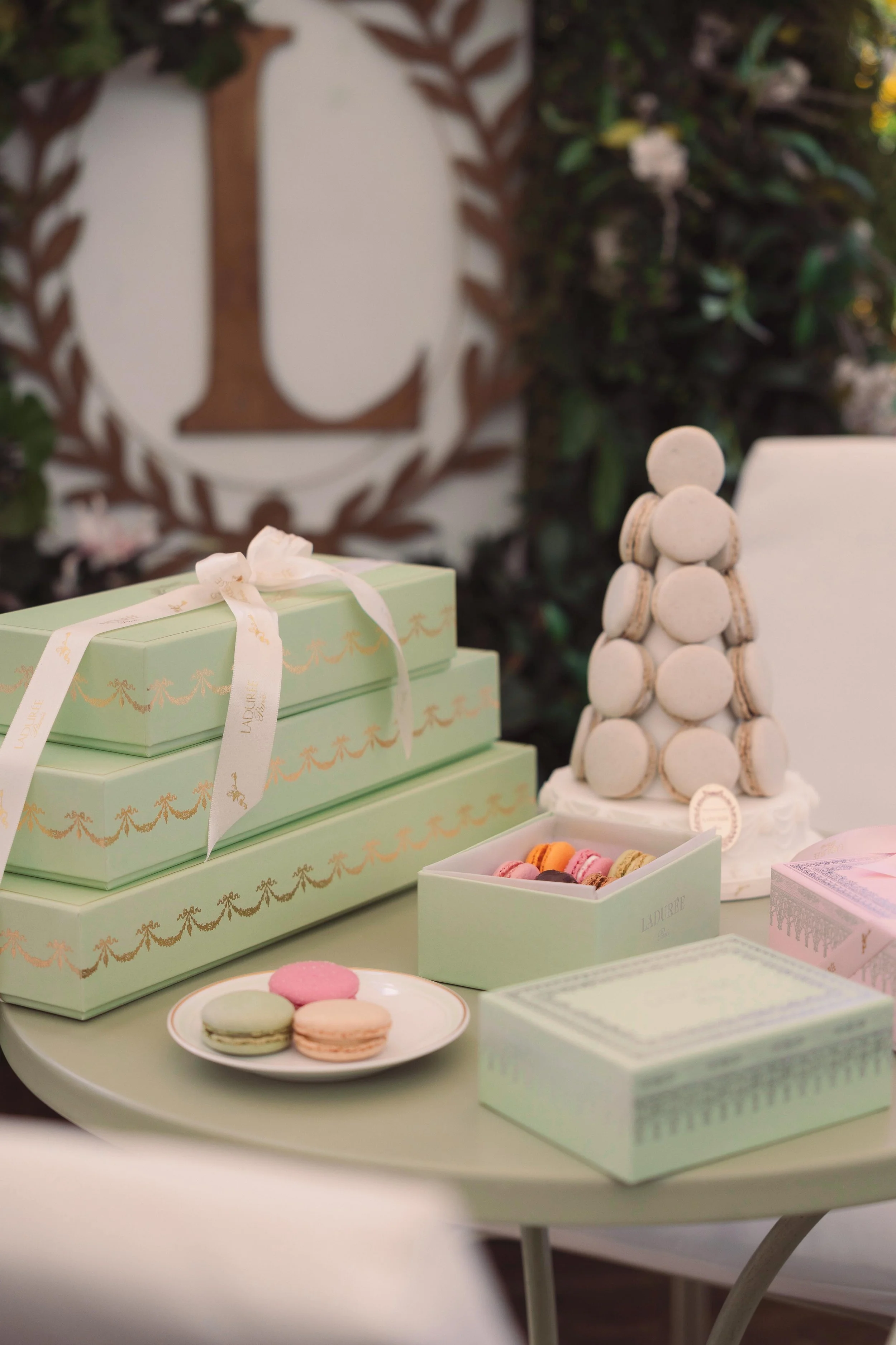 A table displaying pastel-colored gift boxes, macarons, a tower of macarons, and a decorative display in a setting with a wooden logo and greenery in the background.