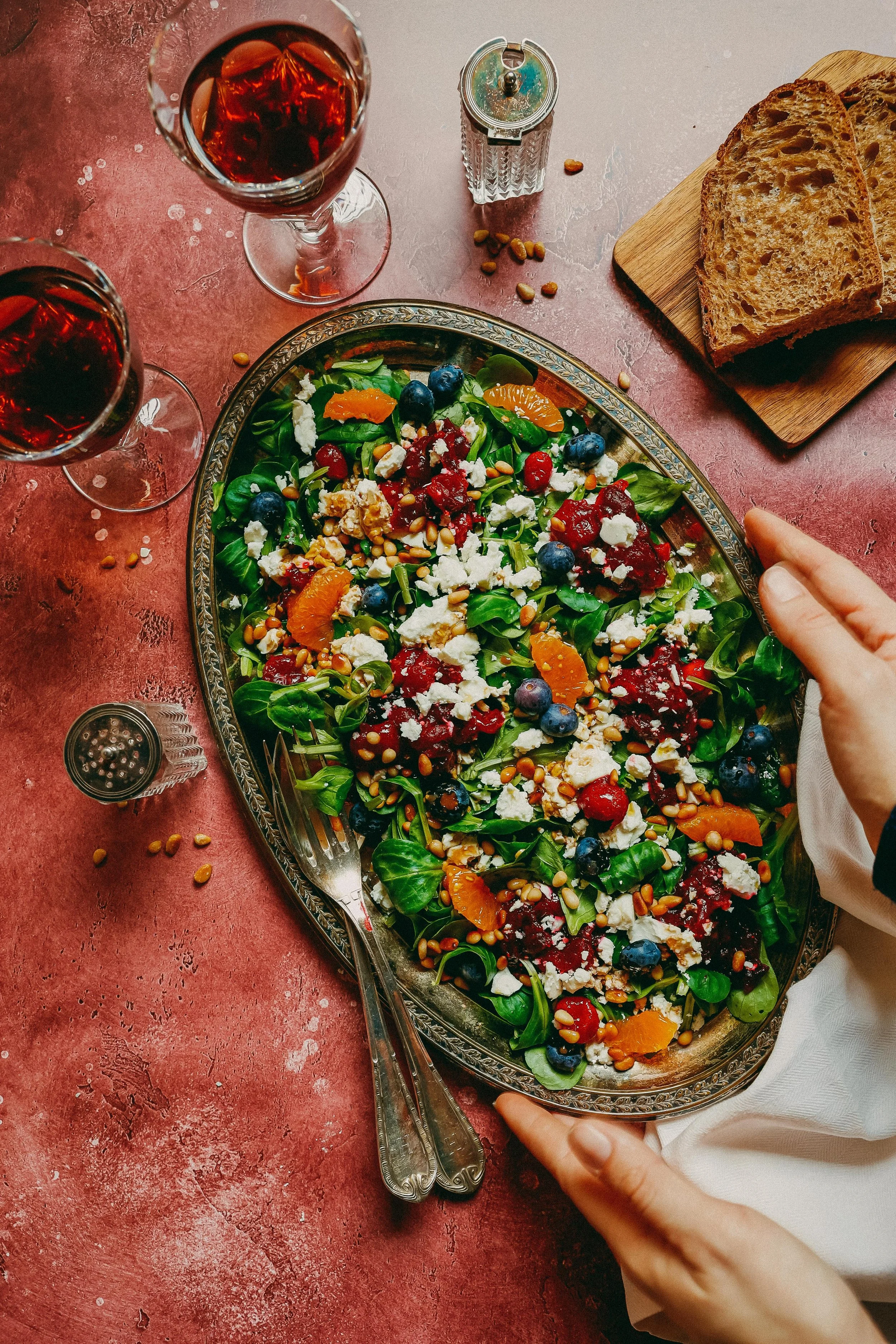 A hand holding a silver tray with a fresh garden salad topped with blueberries, orange segments, crumbled cheese, and pine nuts on a pink table. There are two glasses of red wine, slices of toasted bread on a wooden cutting board, and seasoning containers nearby.