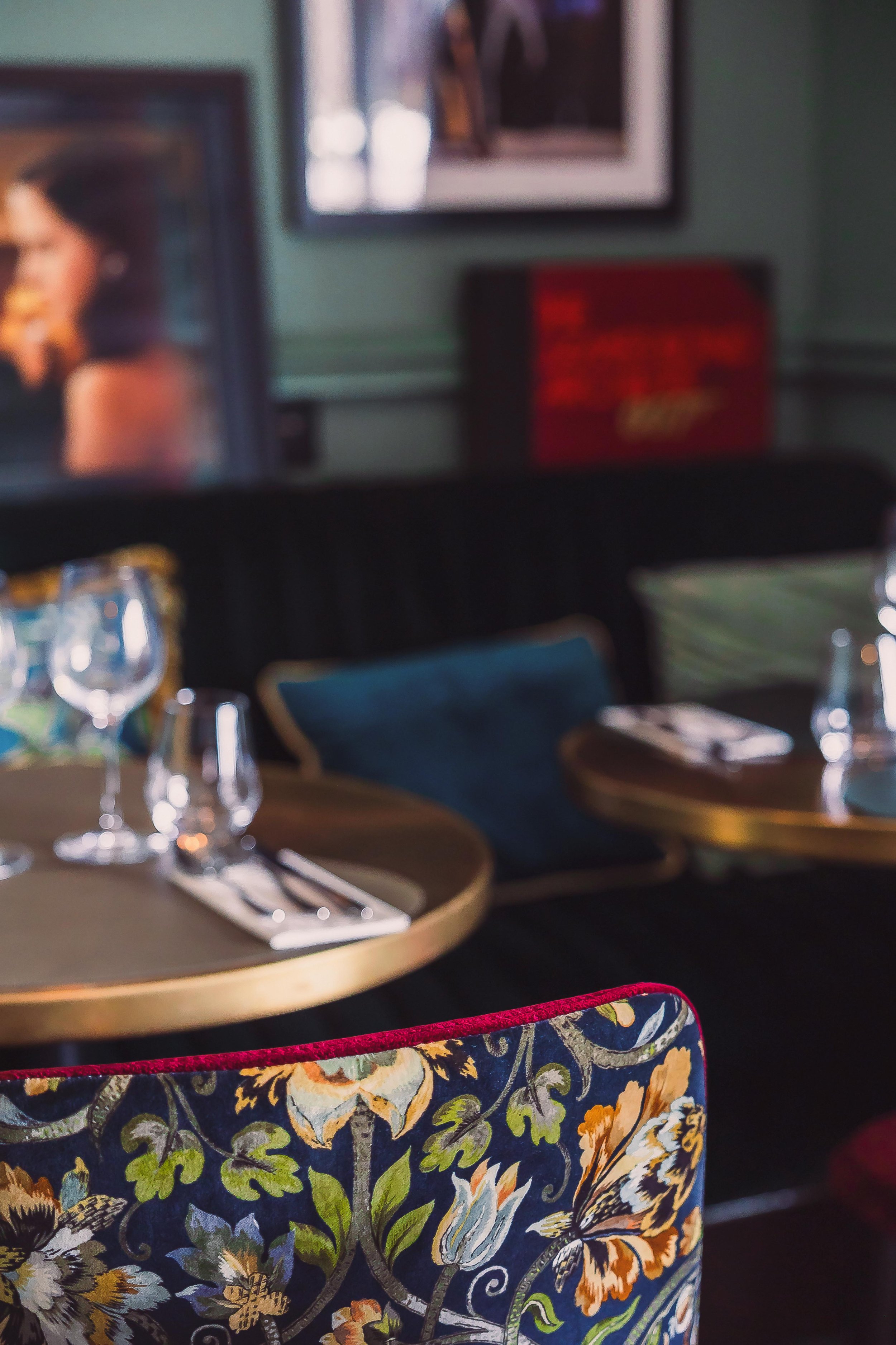Close-up of a brightly patterned floral upholstered chair in a restaurant, with tables set with wine glasses, silverware, and napkins visible in the background.
