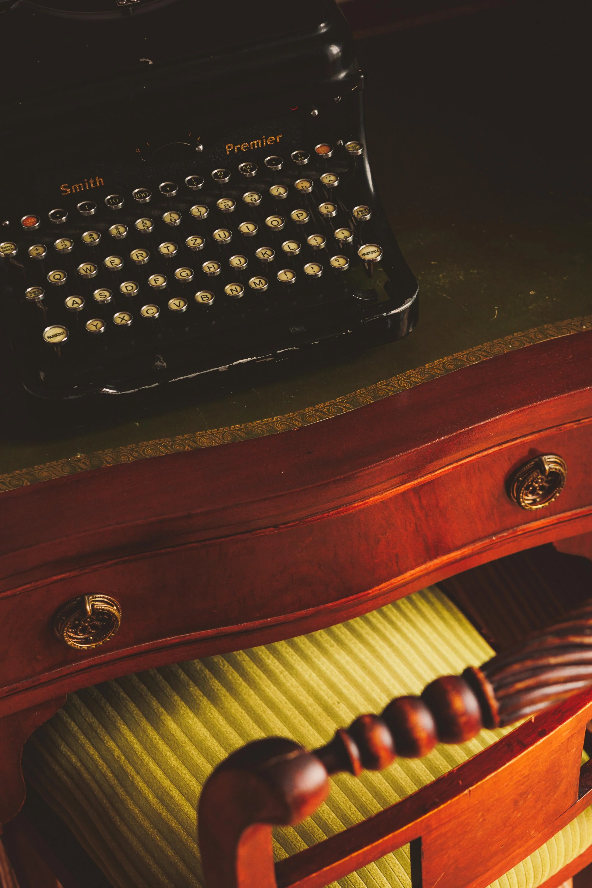 A vintage black typewriter on a wooden desk with a gold trimmed border, and a green upholstered wooden chair beneath the desk.
