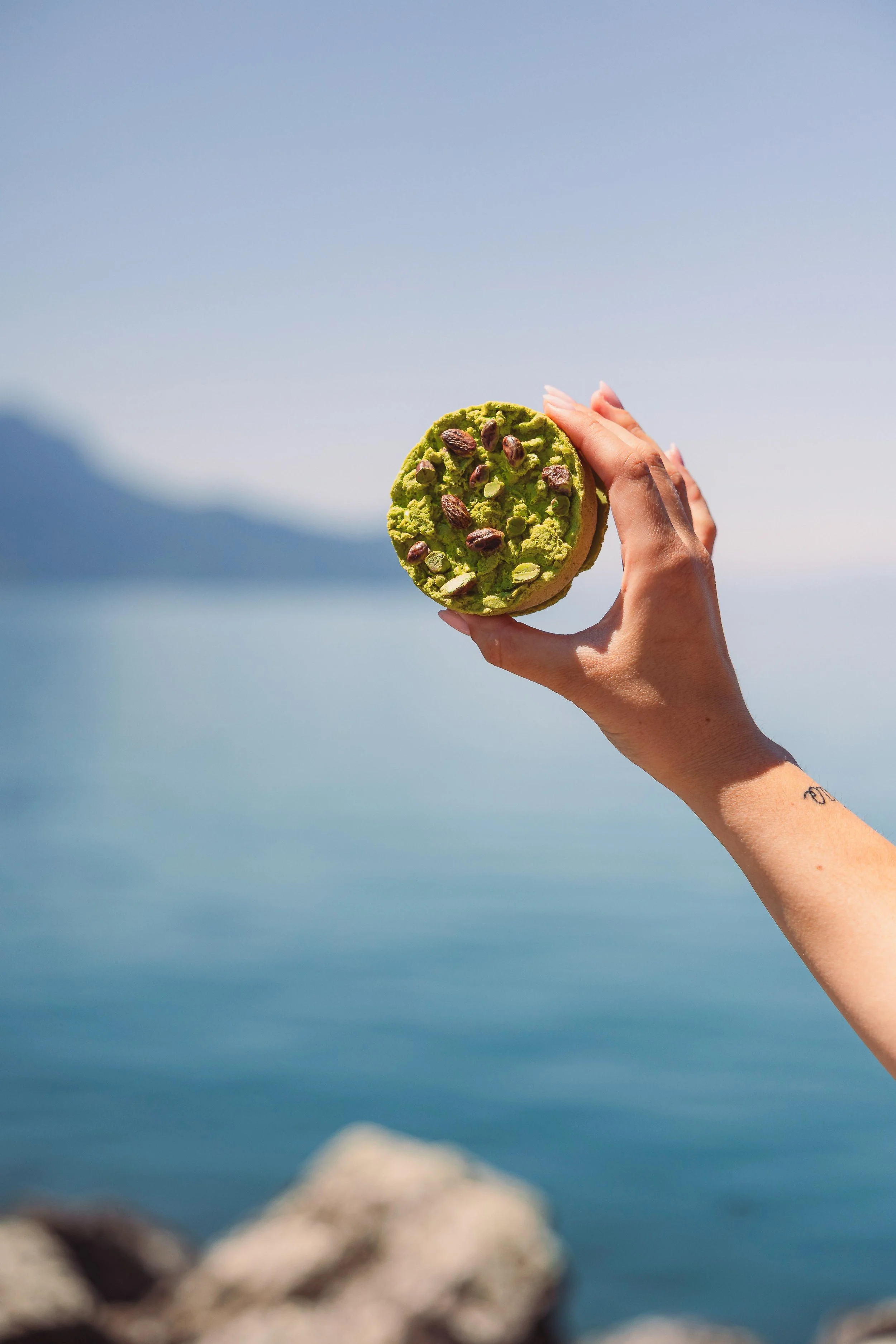 A hand holding a green cookie topped with pistachios against a background of water and mountains.