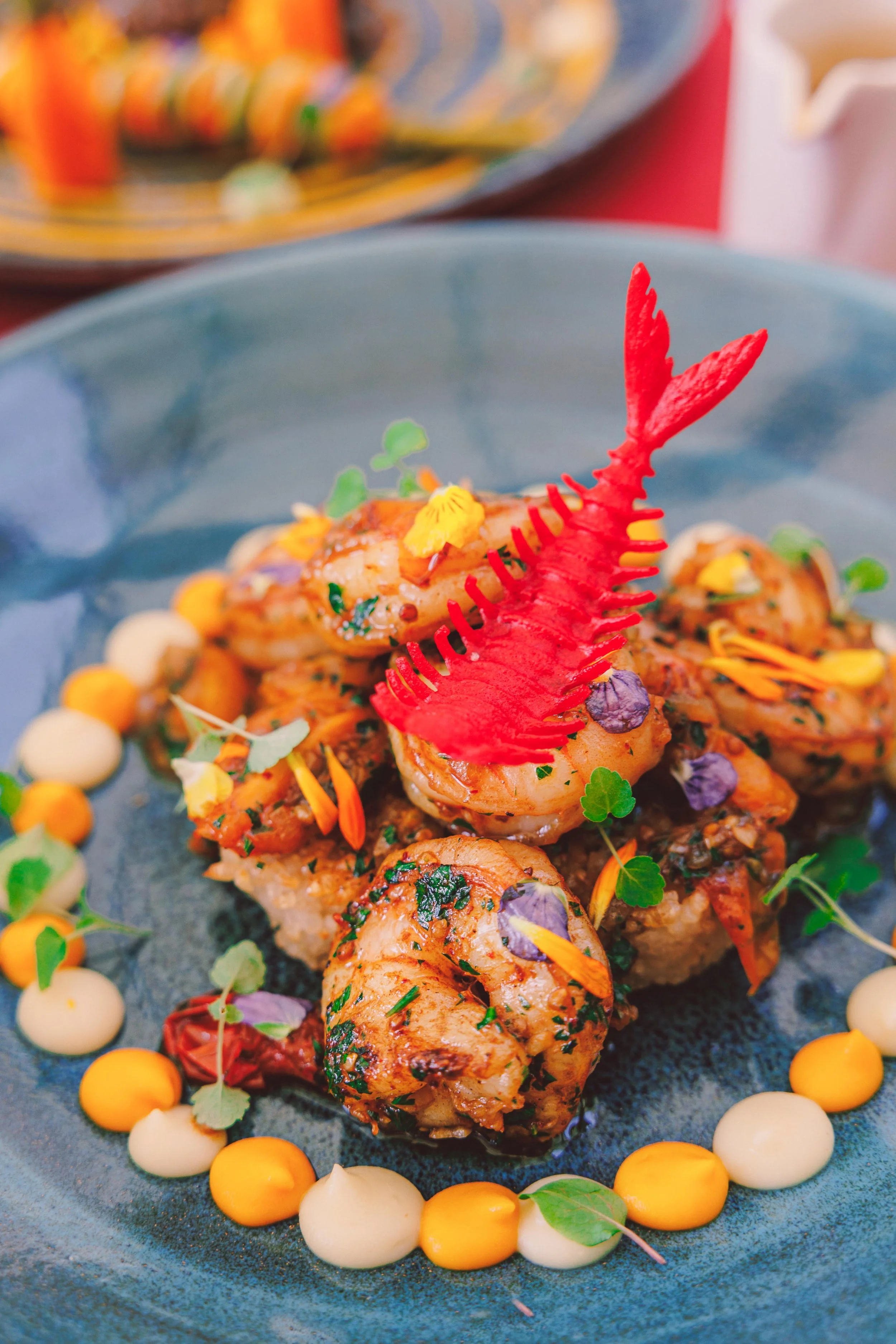 Seafood dish with grilled shrimp, garnished with herbs and edible flowers, served on a black plate with a decorative red lobster-shaped seafood pick, surrounded by yellow and white sauces.
