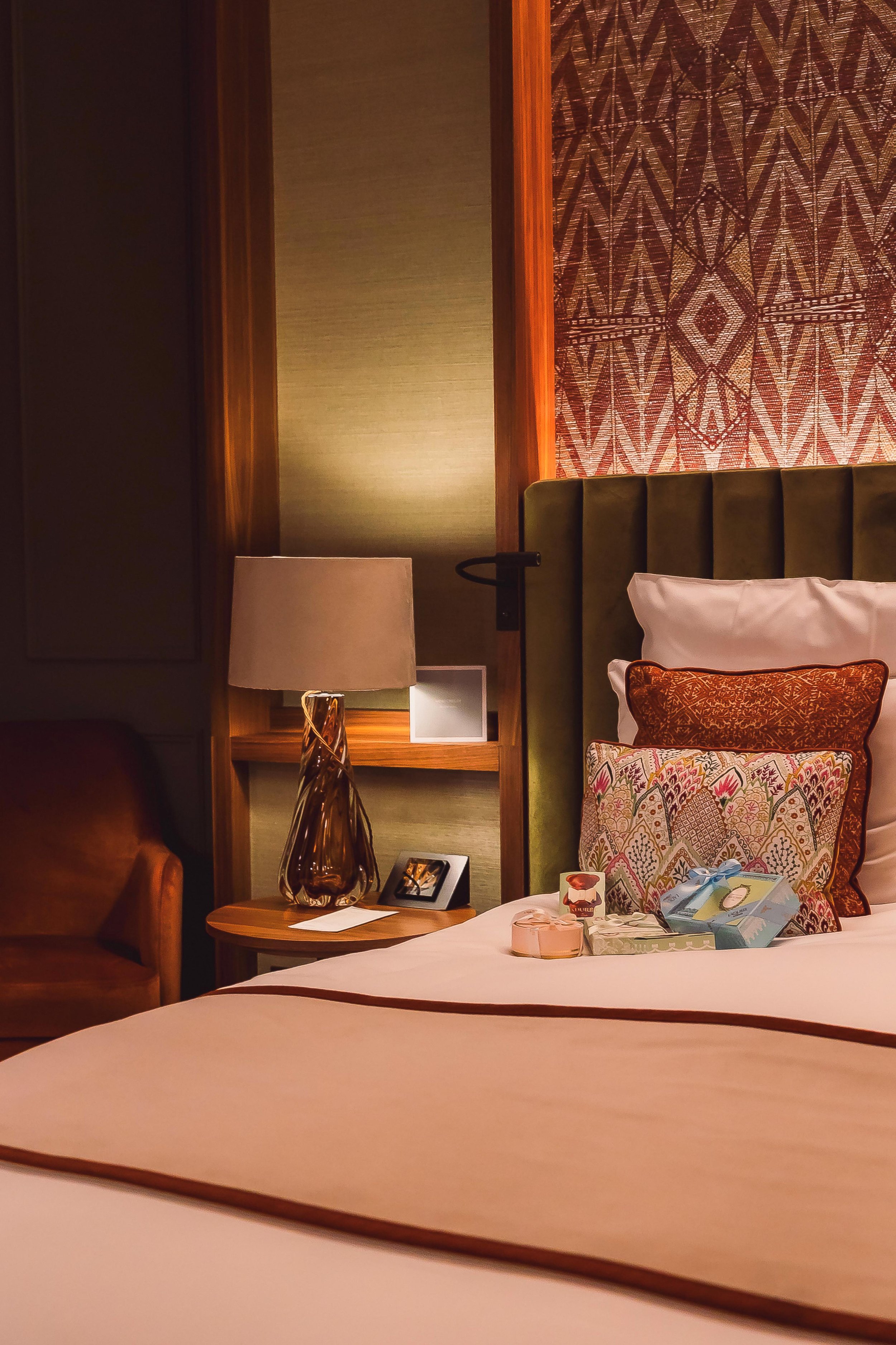 A hotel room with a bed, decorative pillows, and a bedside table with a lamp and photo frame.