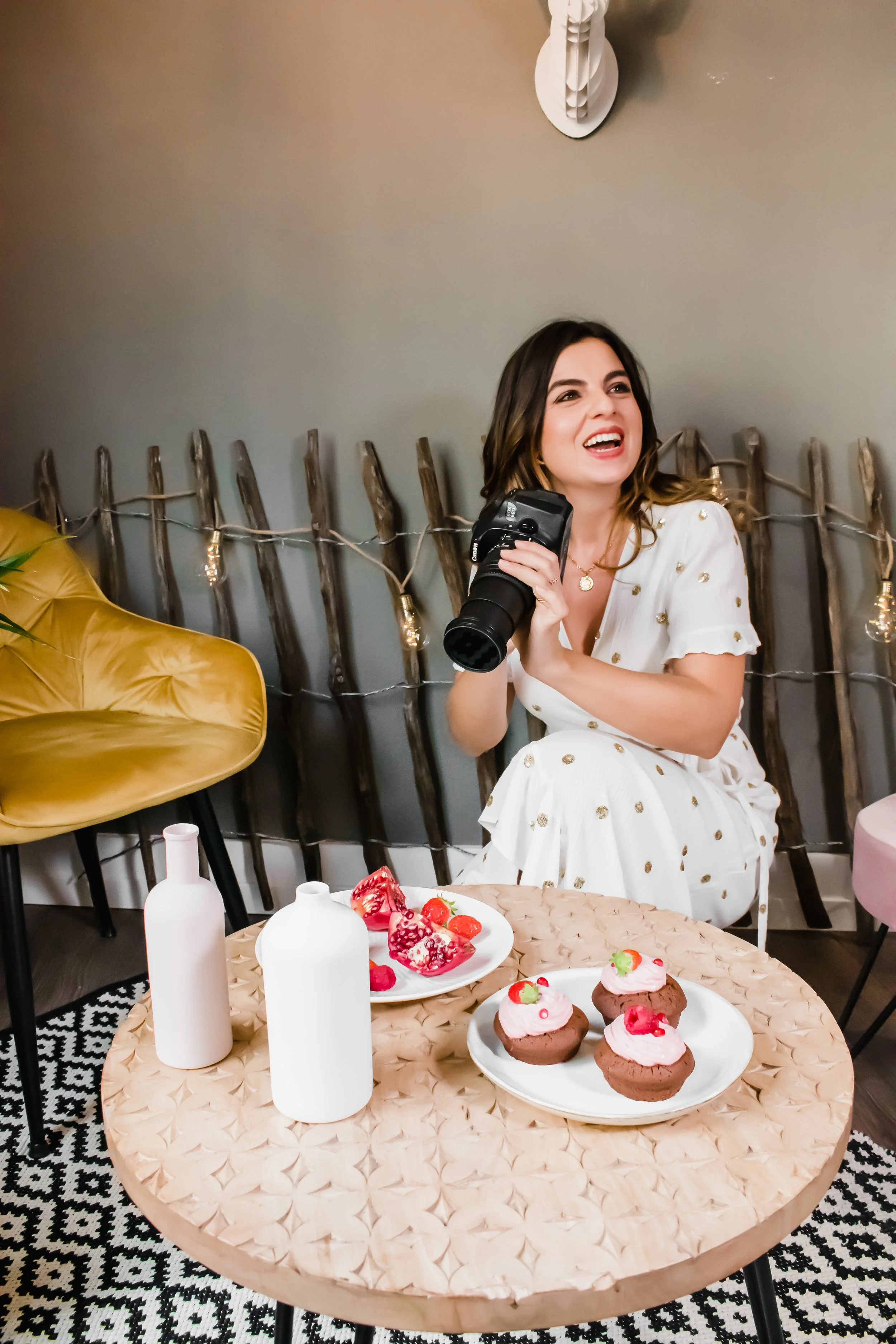 A woman with dark hair in a white dress with gold dots, sitting at a round table with desserts and white bottles, holding a camera and smiling.