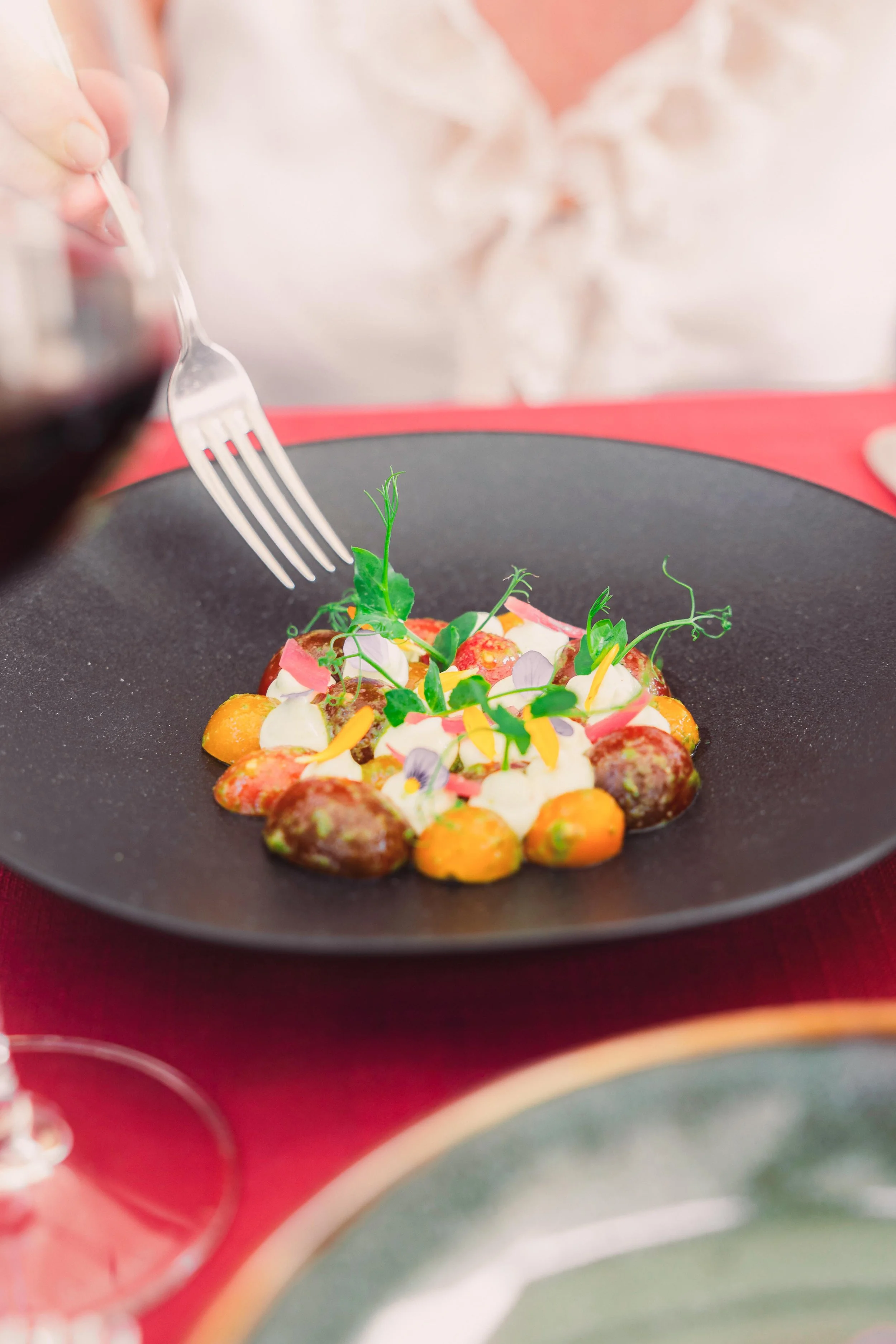 A gourmet dish with colorful vegetables topped with microgreens and edible flowers on a black plate, with a person pouring red wine nearby.