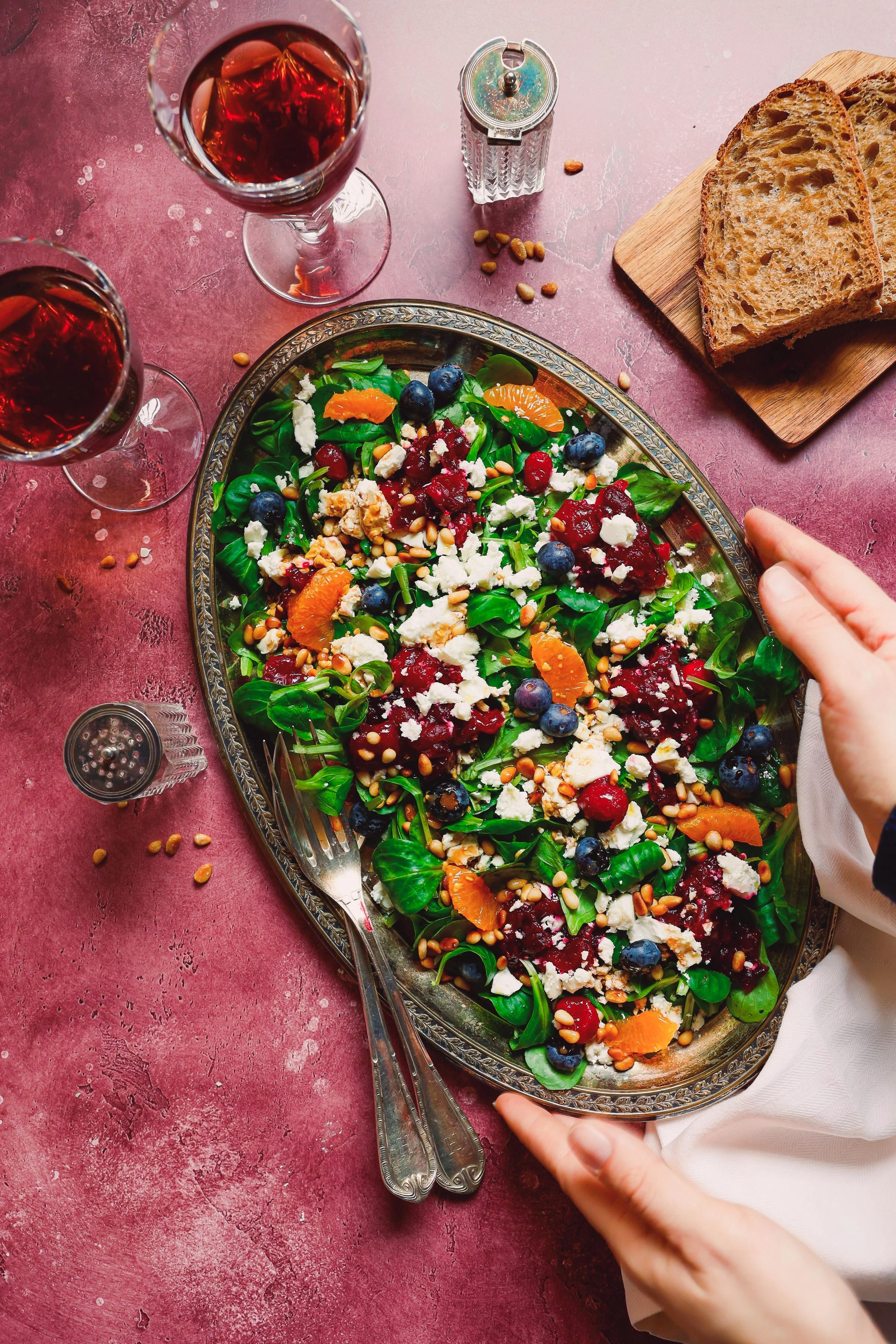 A decorative platter of salad with spinach, blueberries, cranberries, orange slices, crumbled feta cheese, pine nuts, and sunflower seeds on a pink table. Two glasses of red wine, a wooden cutting board with sliced bread, a salt shaker, and a hand re
