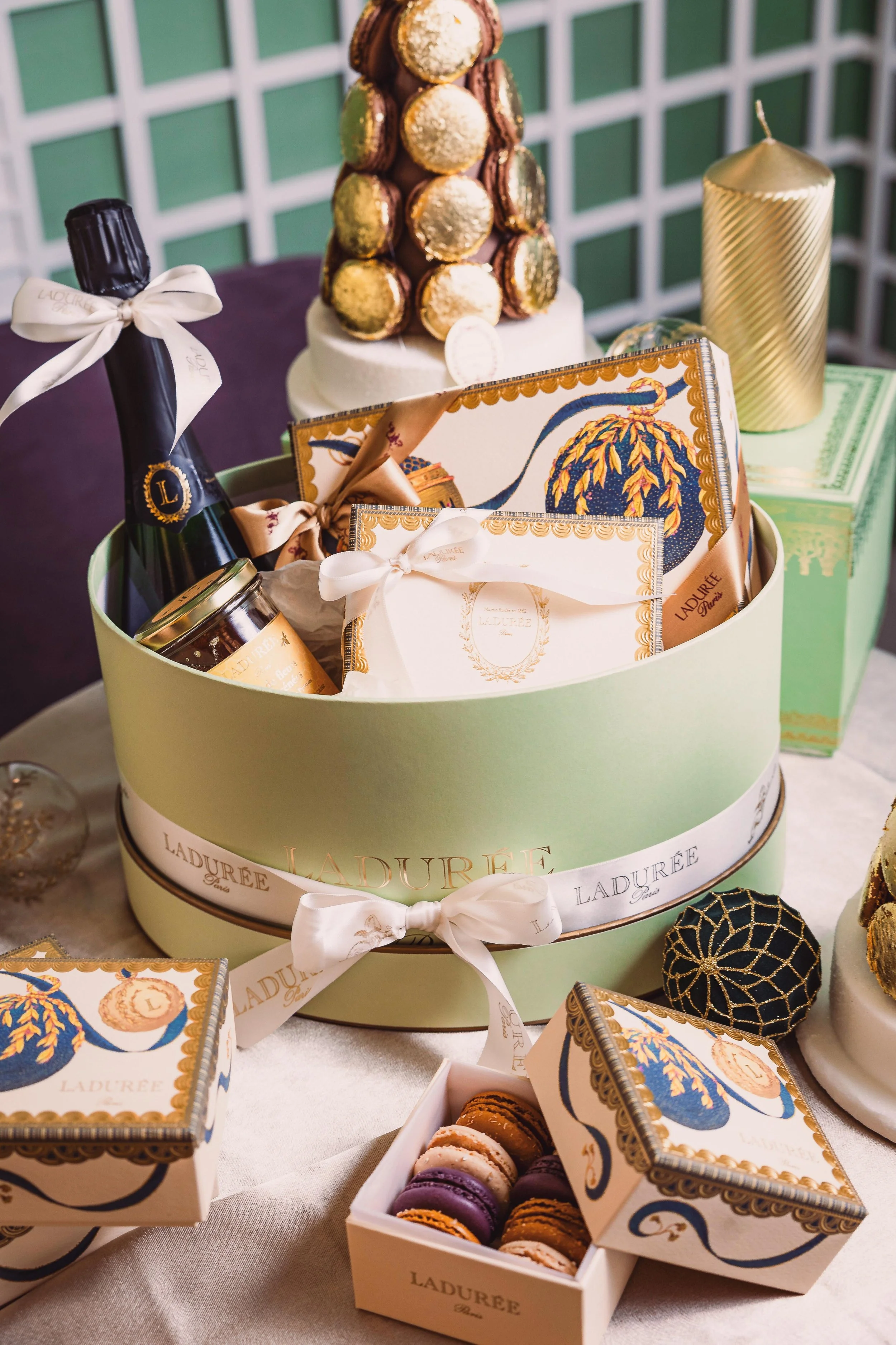 Festive display of Ladurée macarons, a champagne bottle with a white ribbon, a jar of jam, candles, and a decorative box on a table, with a cake topped with macarons in the background.