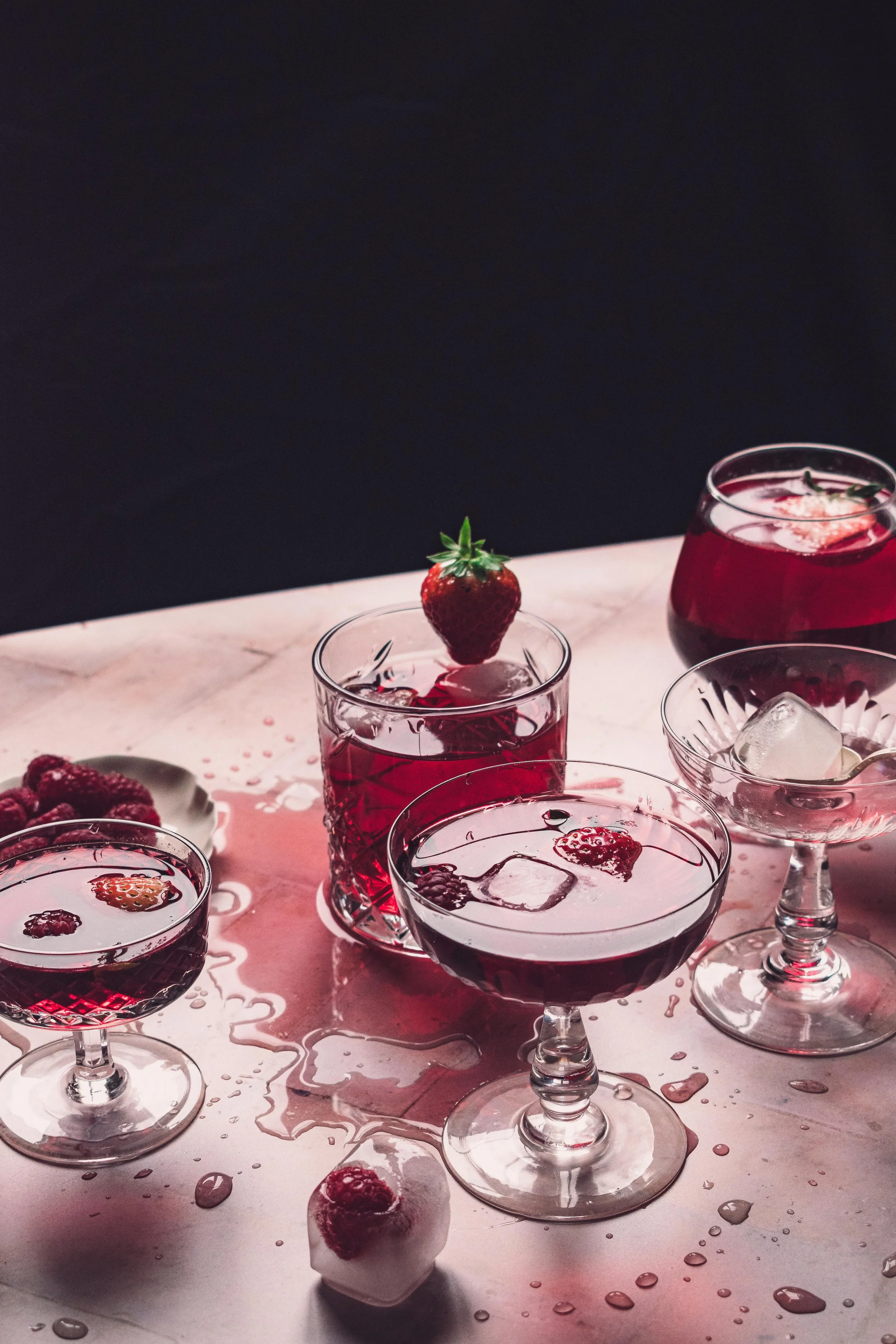 Assorted berry cocktails with strawberries, raspberries, and ice on a pink table with spilled liquid