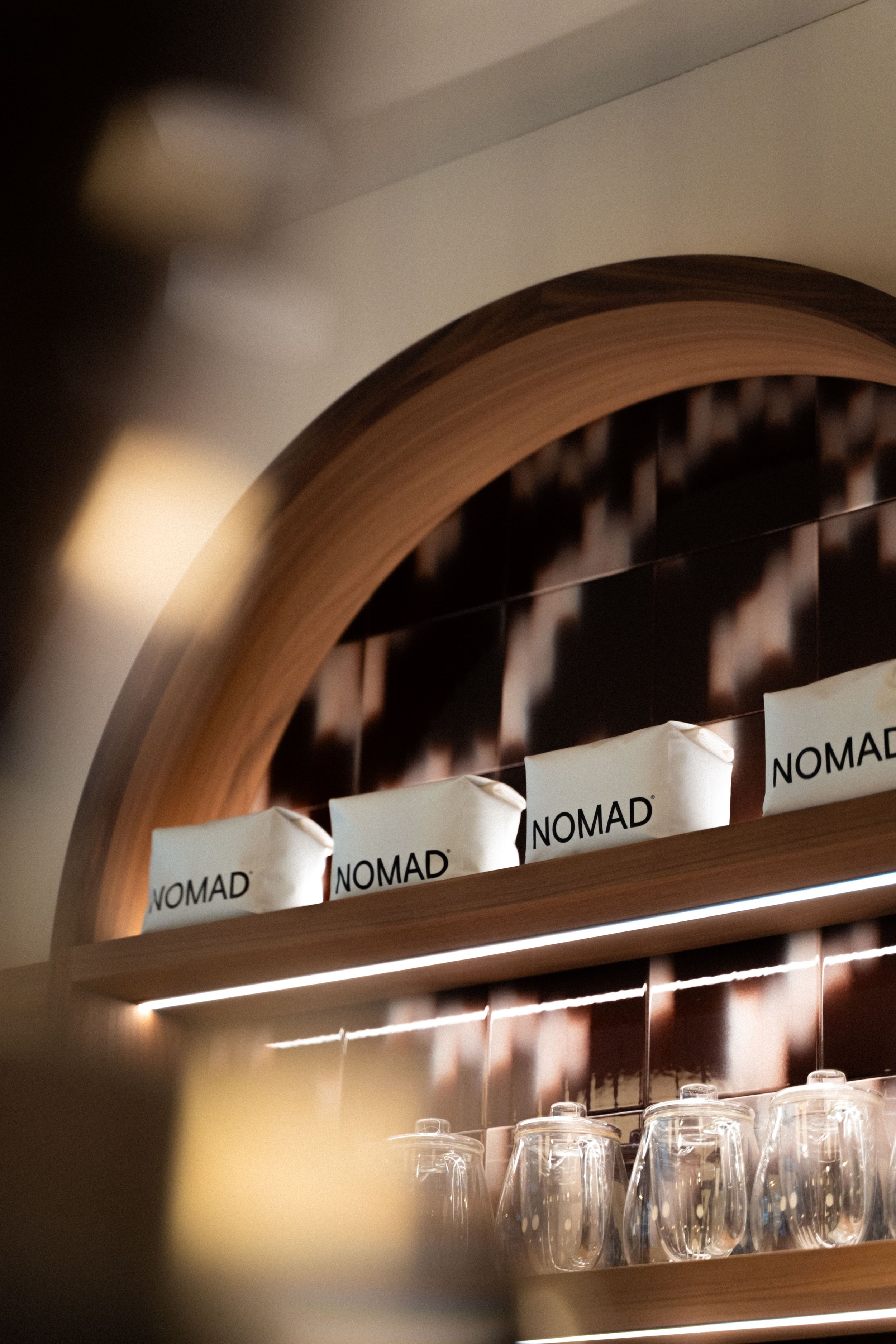 Shelves displaying paper packages labeled 'NOMAD' and upside-down glasses underneath, against a dark tiled wall with a wooden frame.