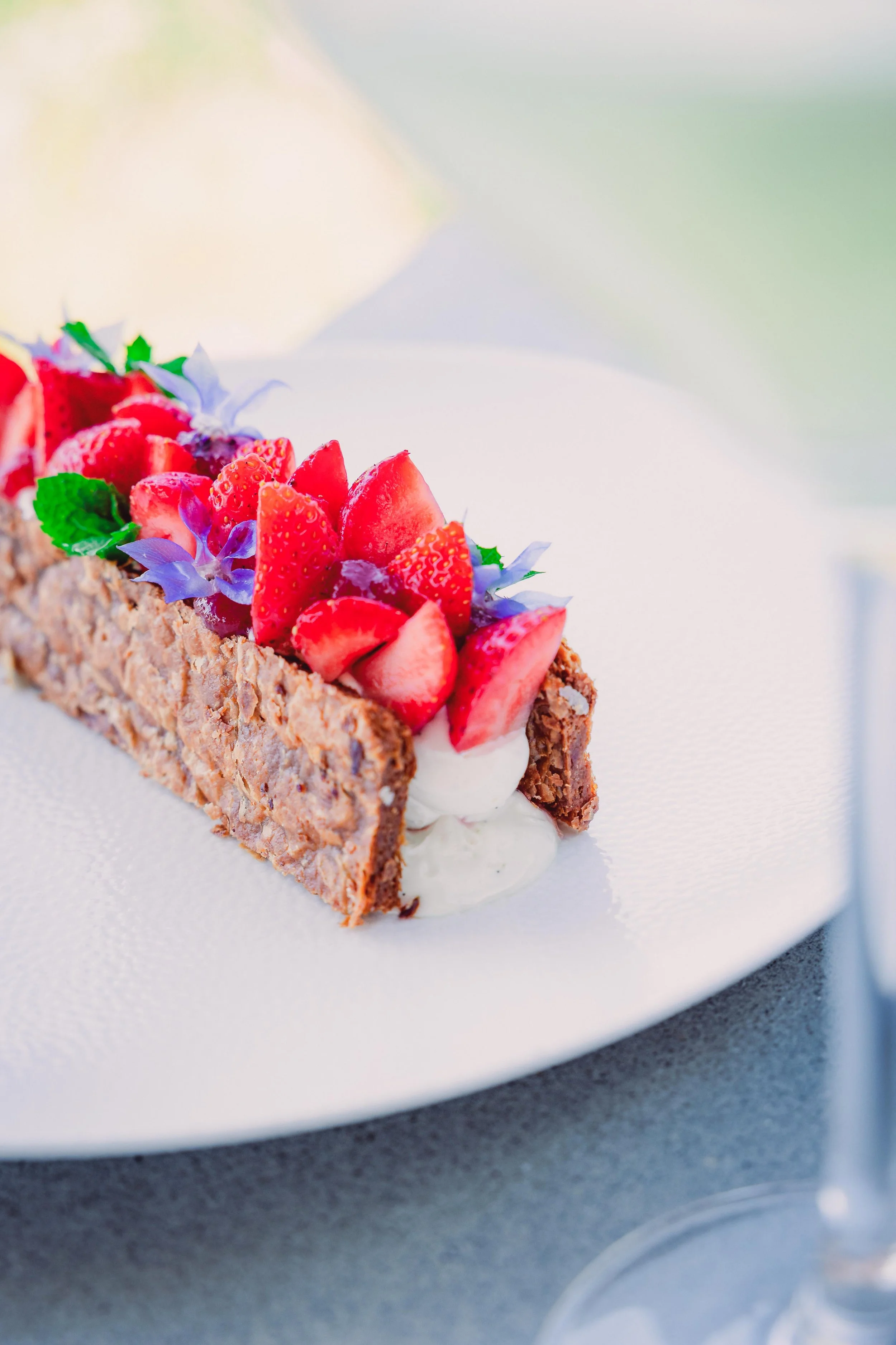 A dessert tart garnished with sliced strawberries, purple flowers, green mint leaves, and a cream filling on a white plate.