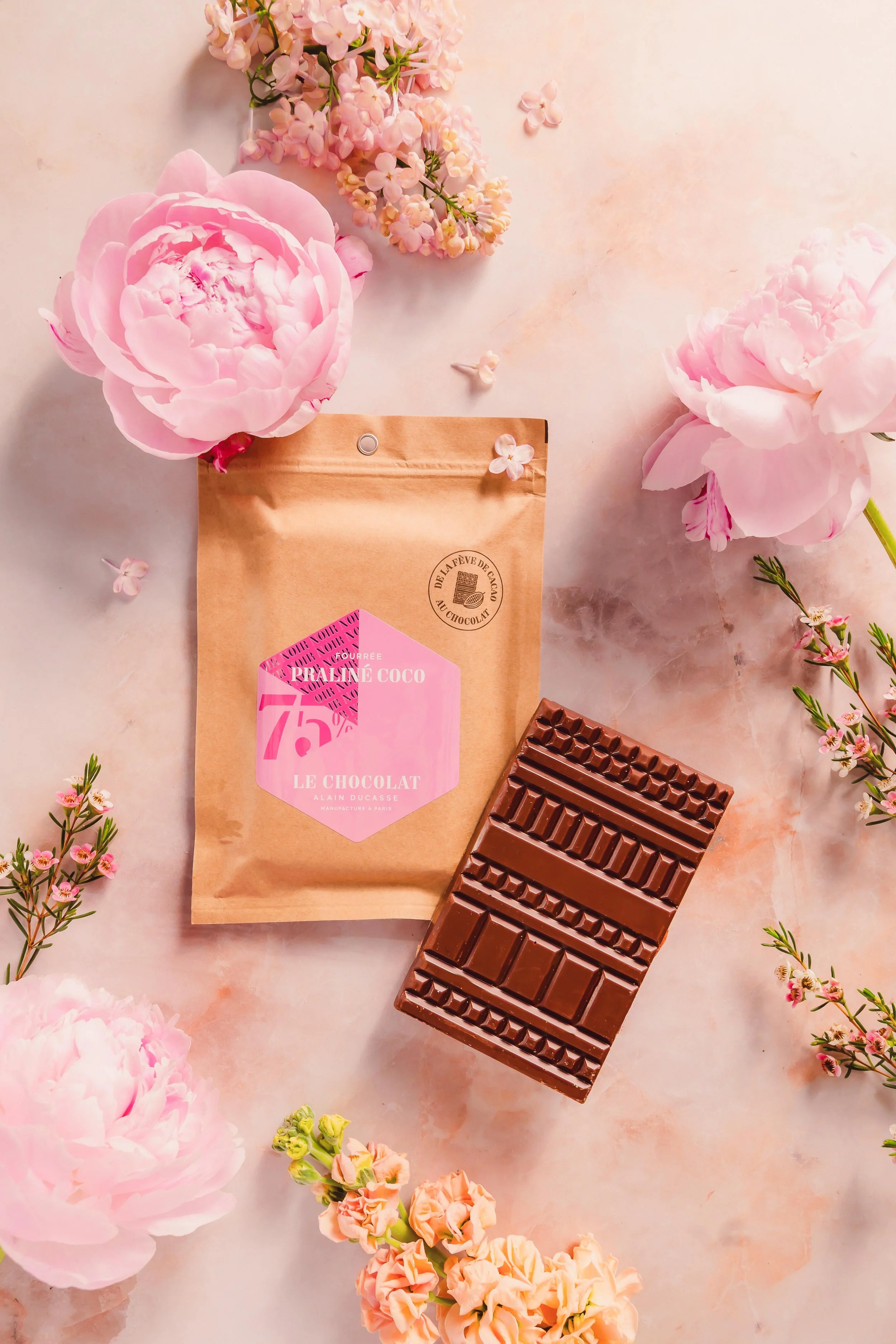 A pink package of praline chocolate from La Fève de Cacao surrounded by pink peonies and small pink and orange flowers on a light pink marble surface.