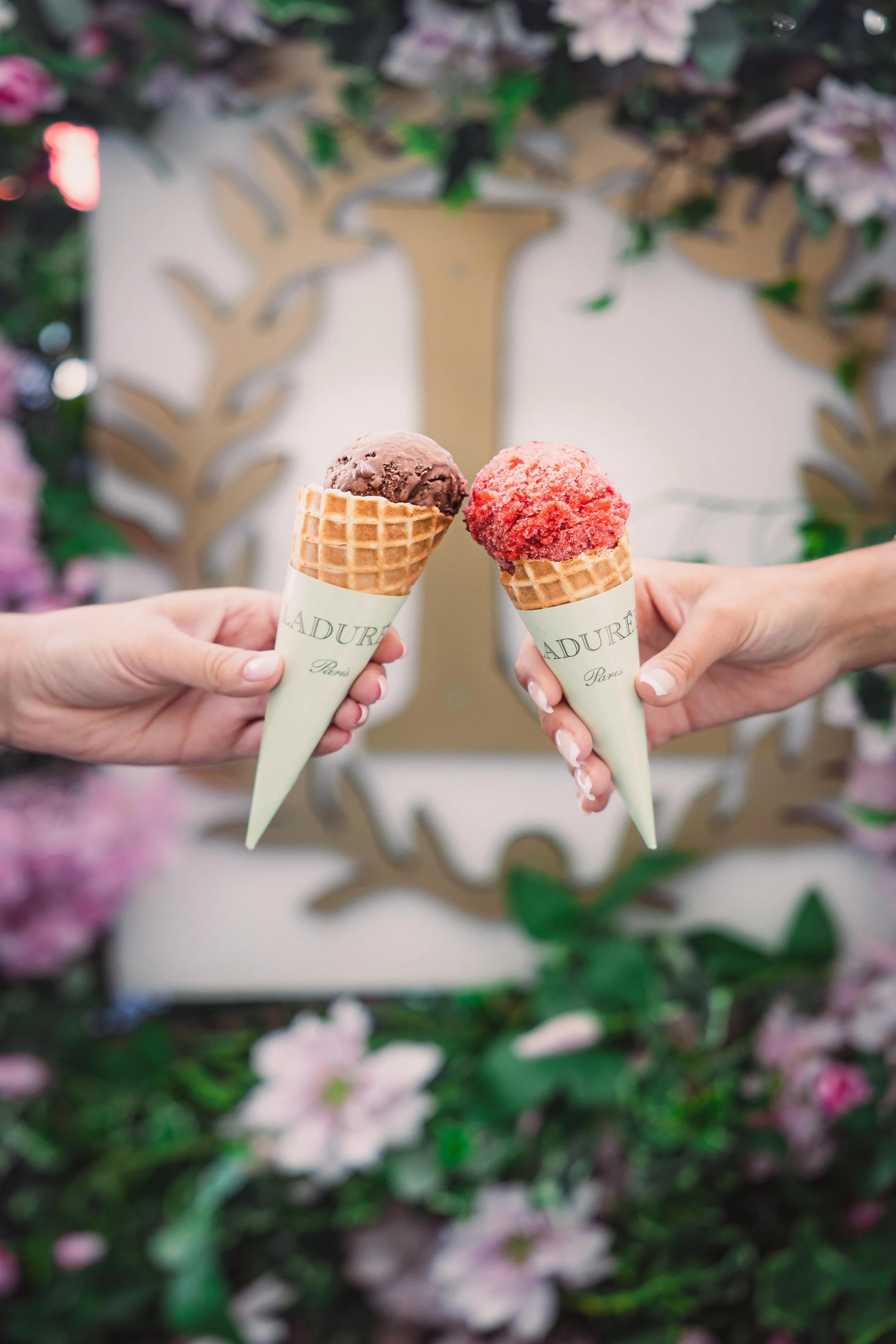 Two hands holding ice cream cones with chocolate and strawberry flavored scoops, background with floral decorations.