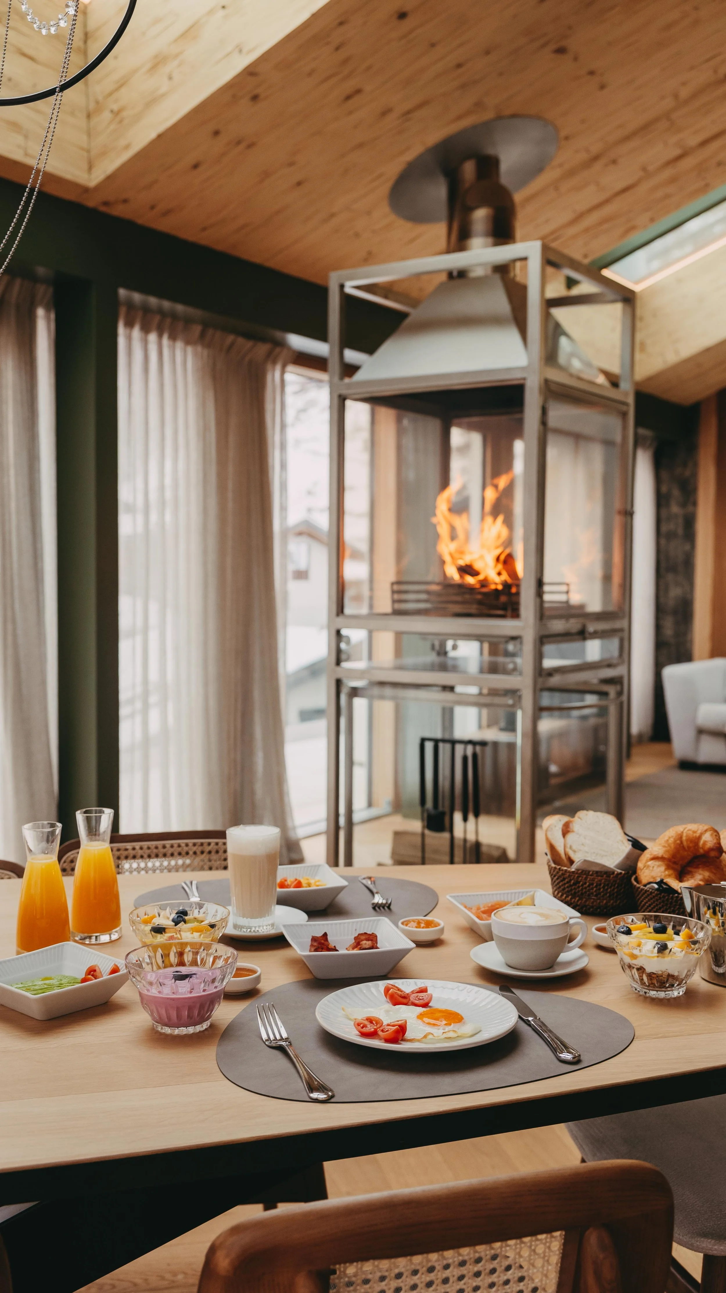 A breakfast spread on a wooden dining table with various dishes, drinks, and a fireplace in the background inside a modern home.
