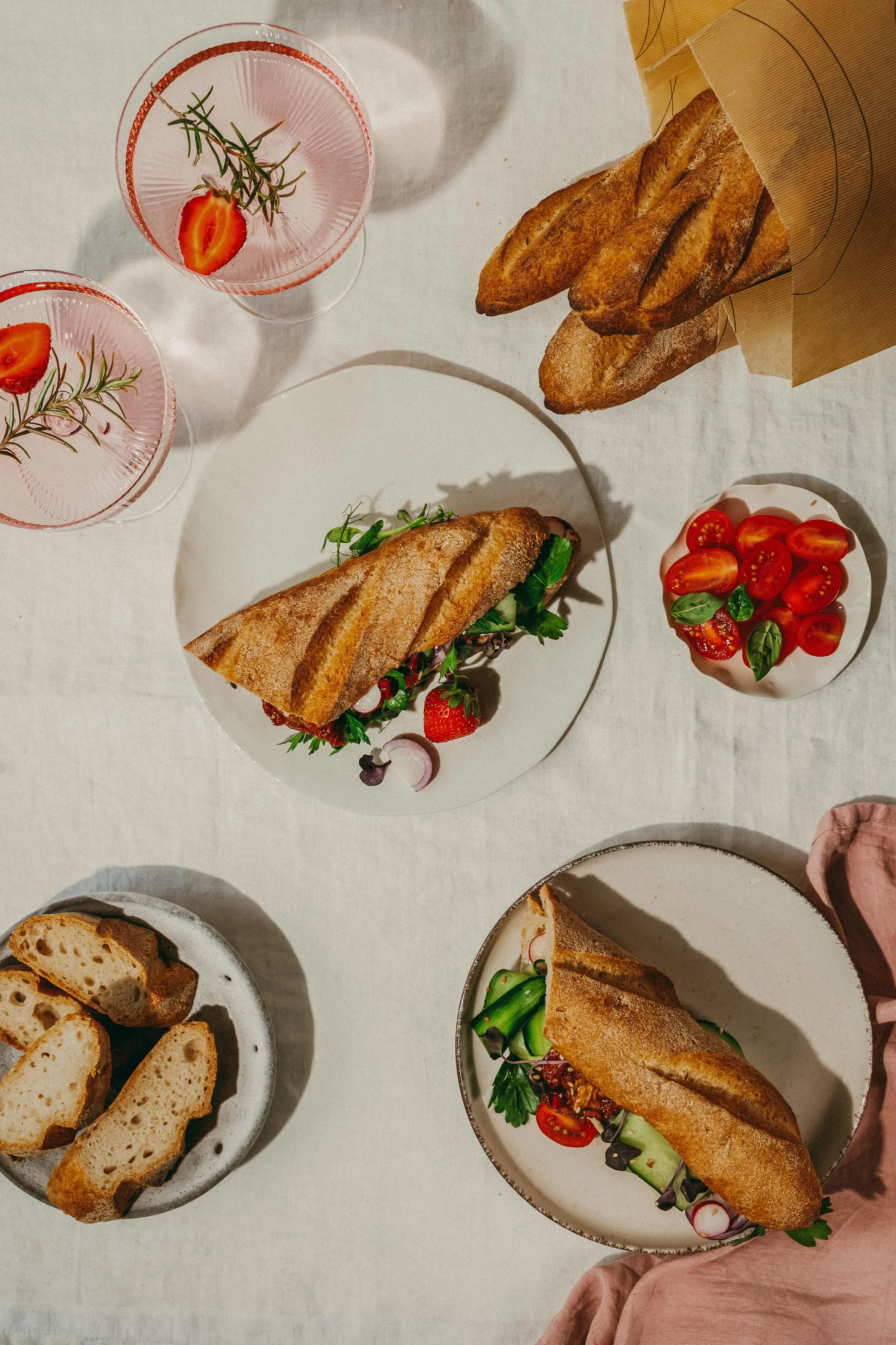 A table set with bread, sandwiches, cherry tomatoes, and drinks garnished with herbs.