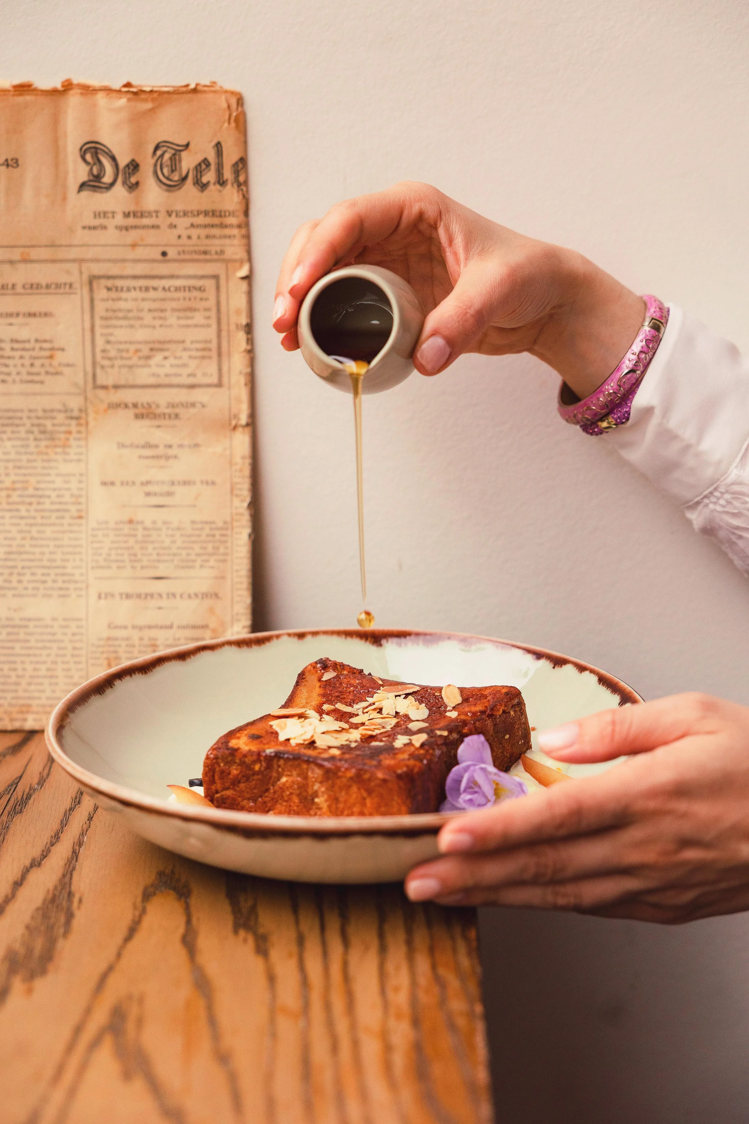 Hand pouring syrup over a rectangular slice of French toast garnished with sliced almonds and purple flowers on a white plate, with a vintage newspaper in the background.