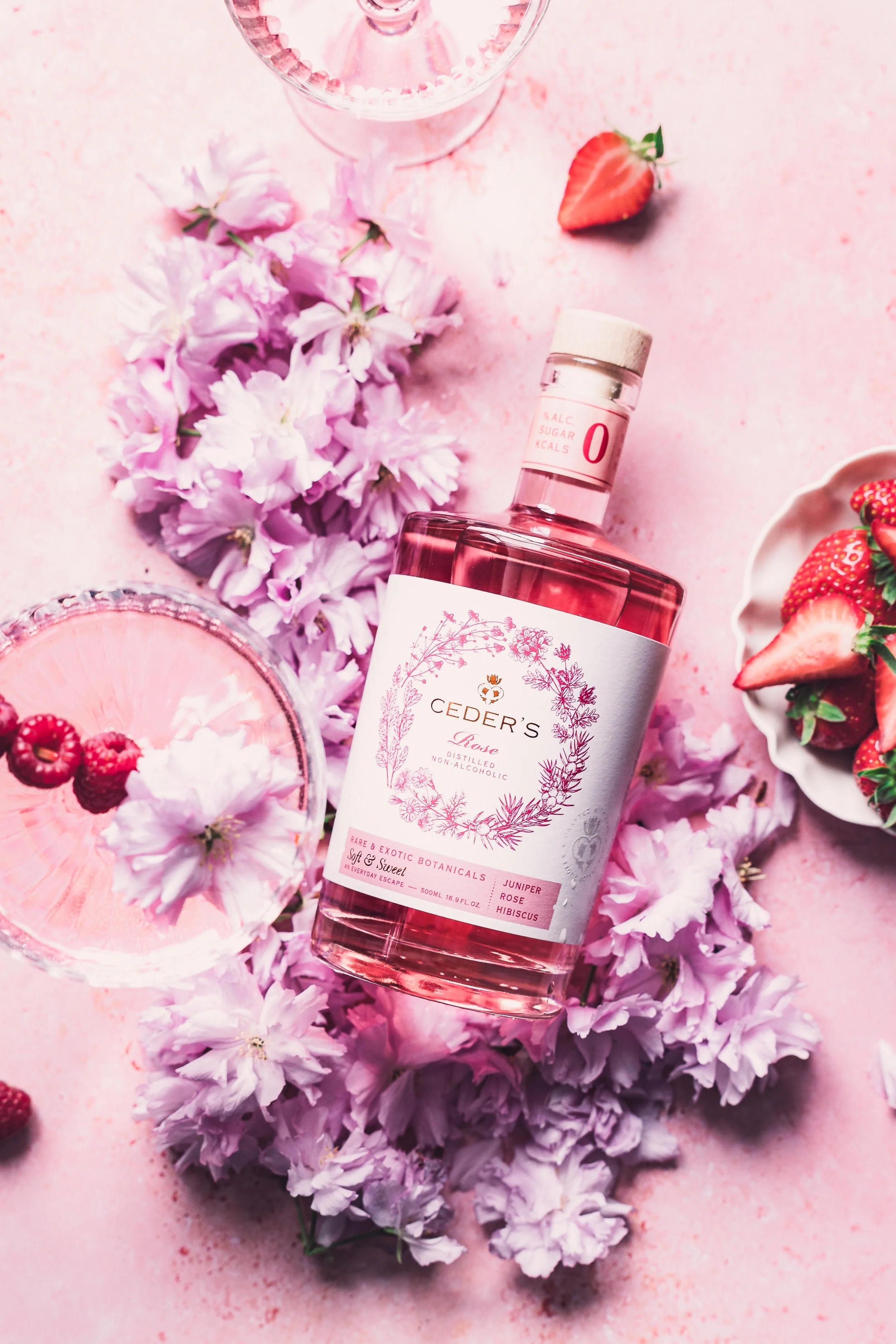 A bottle of Ceder's Rose non-alcoholic spirit surrounded by pink flowers, strawberries, and raspberry garnishes on a pink background.