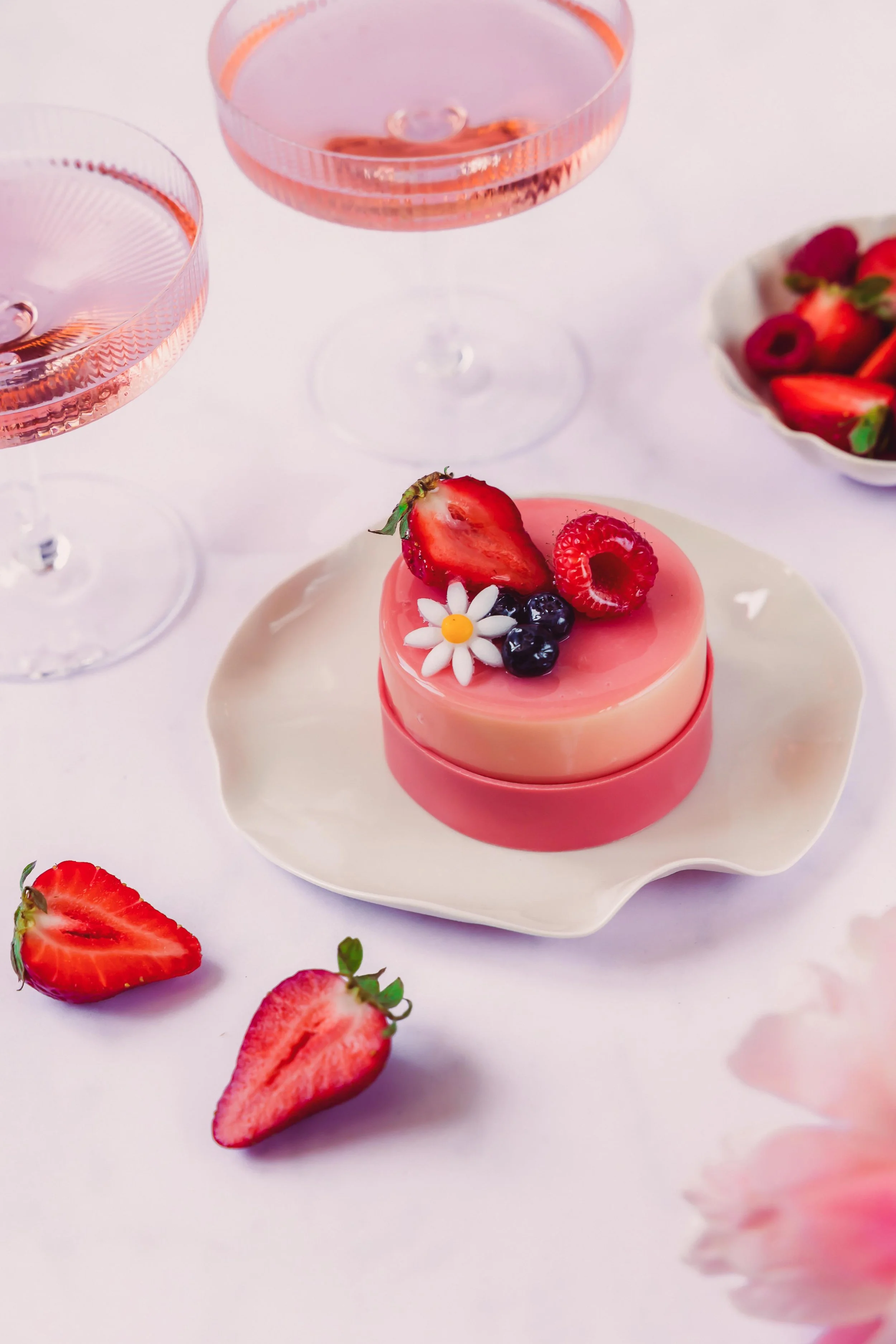 A small round pink and white dessert topped with fresh strawberries, raspberries, blueberries, and a white daisy-shaped edible decoration on a decorative white plate; two glasses of pink bubbly drink and a bowl of strawberries in the background.