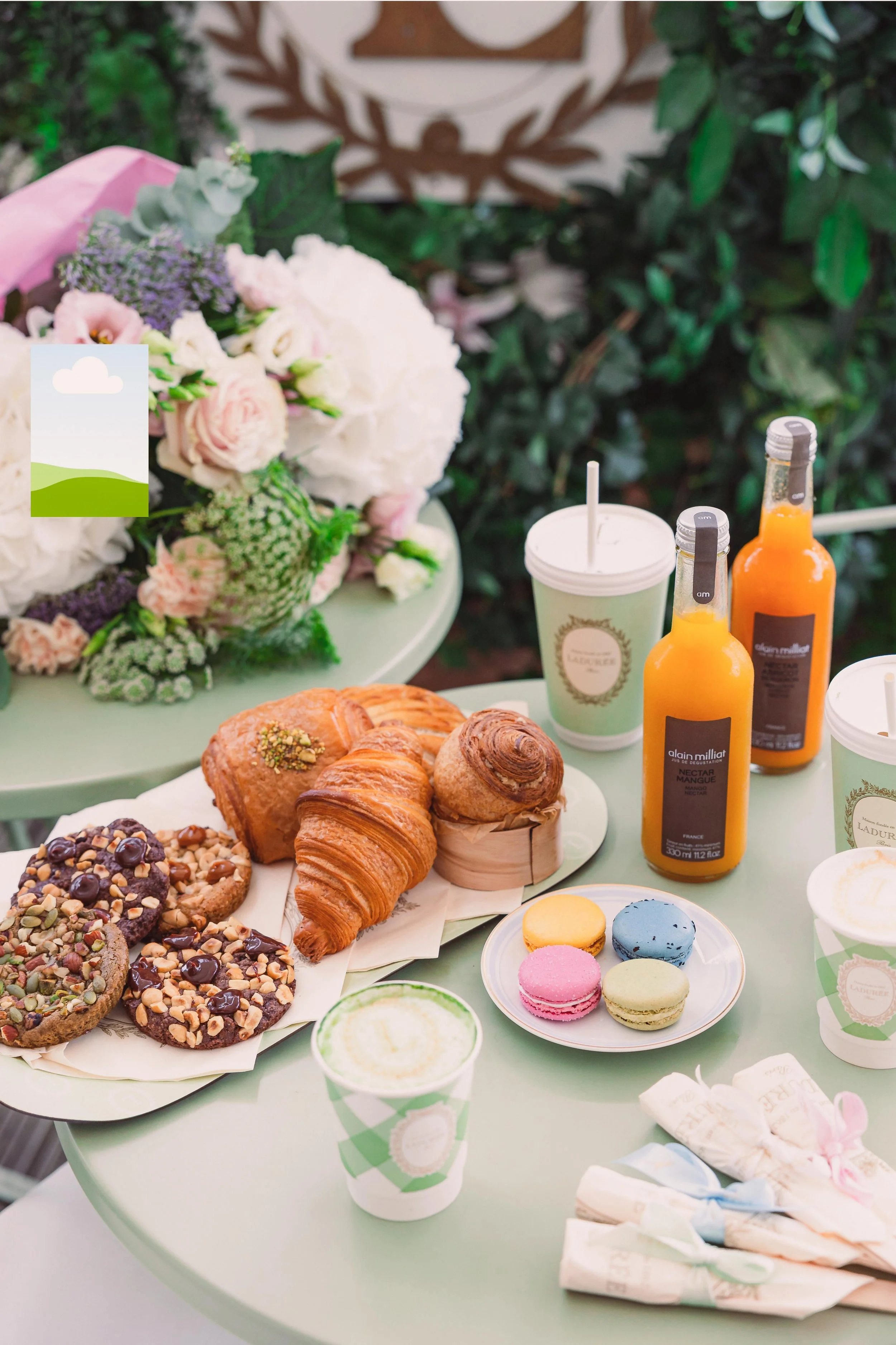 A pastel green table with assorted baked goods, including croissants, cookies topped with nuts and chocolate chips, and macarons in pink, yellow, green, and blue. Two bottles of nectar mango juice, drink cups, and a floral bouquet with white and pink