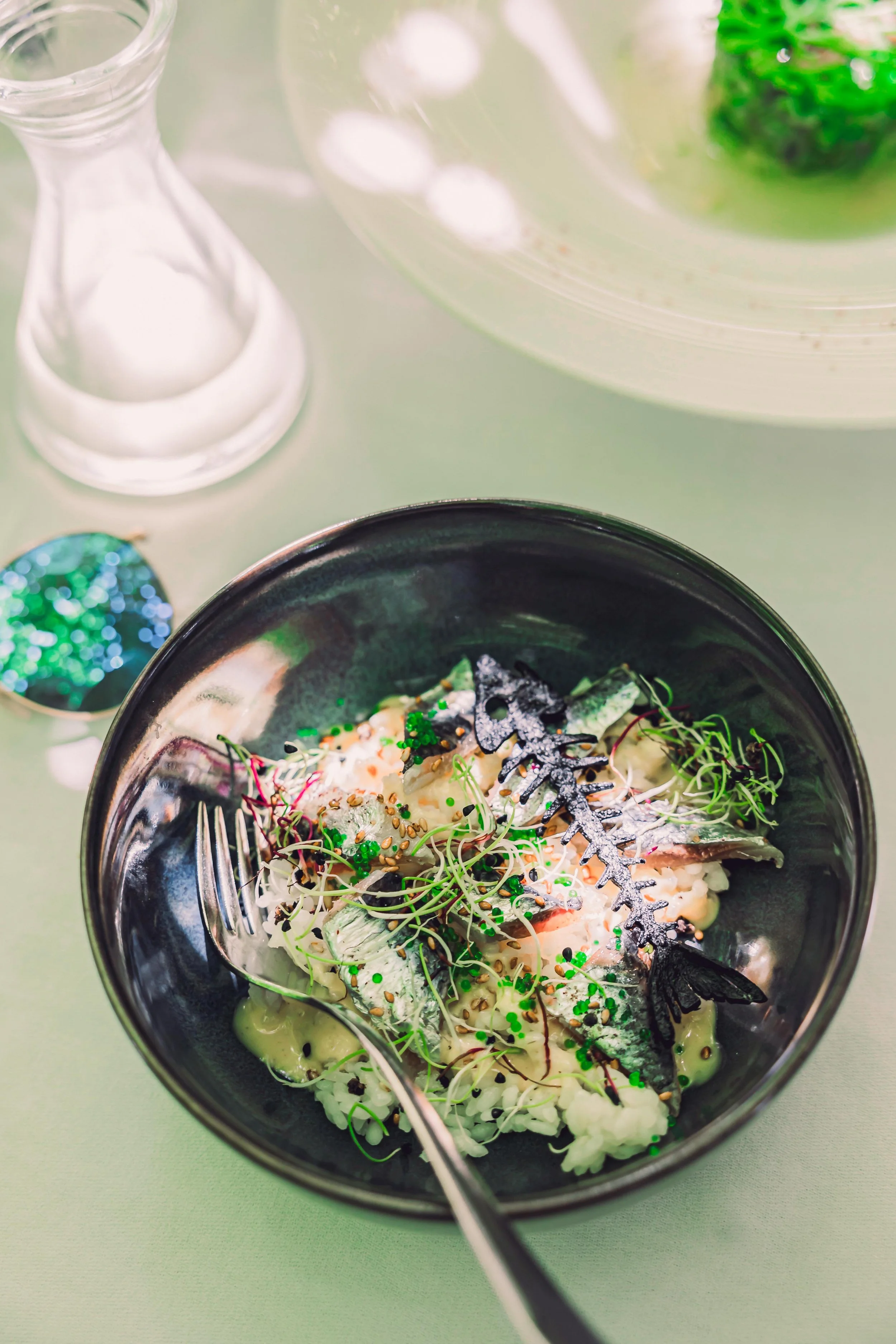 A salad with microgreens, chopped fish, and sauce in a dark bowl on a table, with a glass bottle and decorative object nearby.