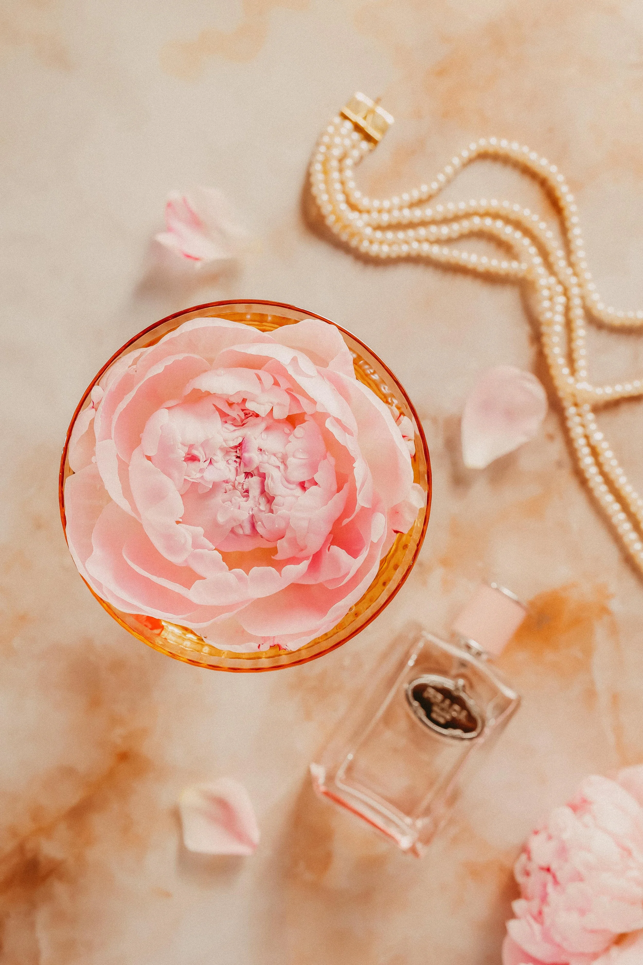 Top view of a pink peony flower-shaped candle in a glass holder, scattered pink flower petals, a pearl necklace, and a perfume bottle on a beige surface.